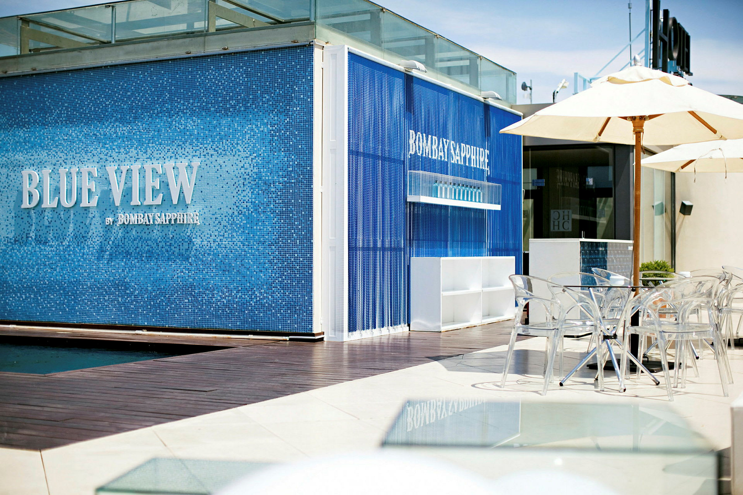 En moderne bar facadedesign med blå fliser og teksten "Blue View by Bombay Sapphire". Foran er gennemsigtige stole og parasoller. (AI-genereret tekst)