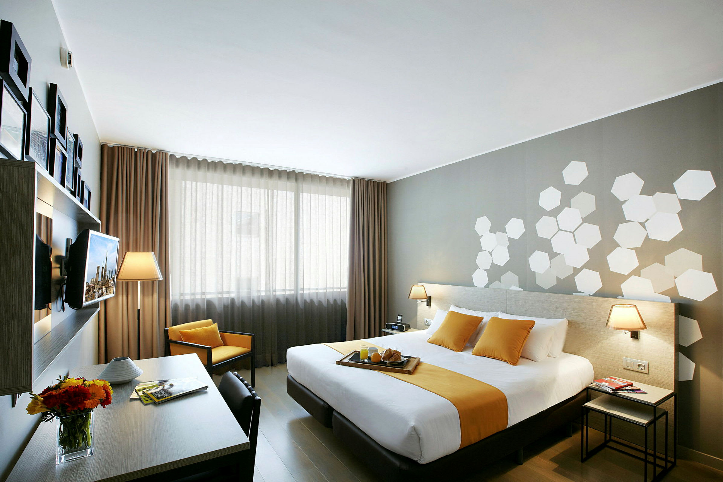 Et moderne hotelværelse med en dobbeltseng dækket af hvidt og gult sengetøj. I rummet er der et skrivebord med en blomsterkrukke og lamper på natbordene. (AI-genereret tekst)