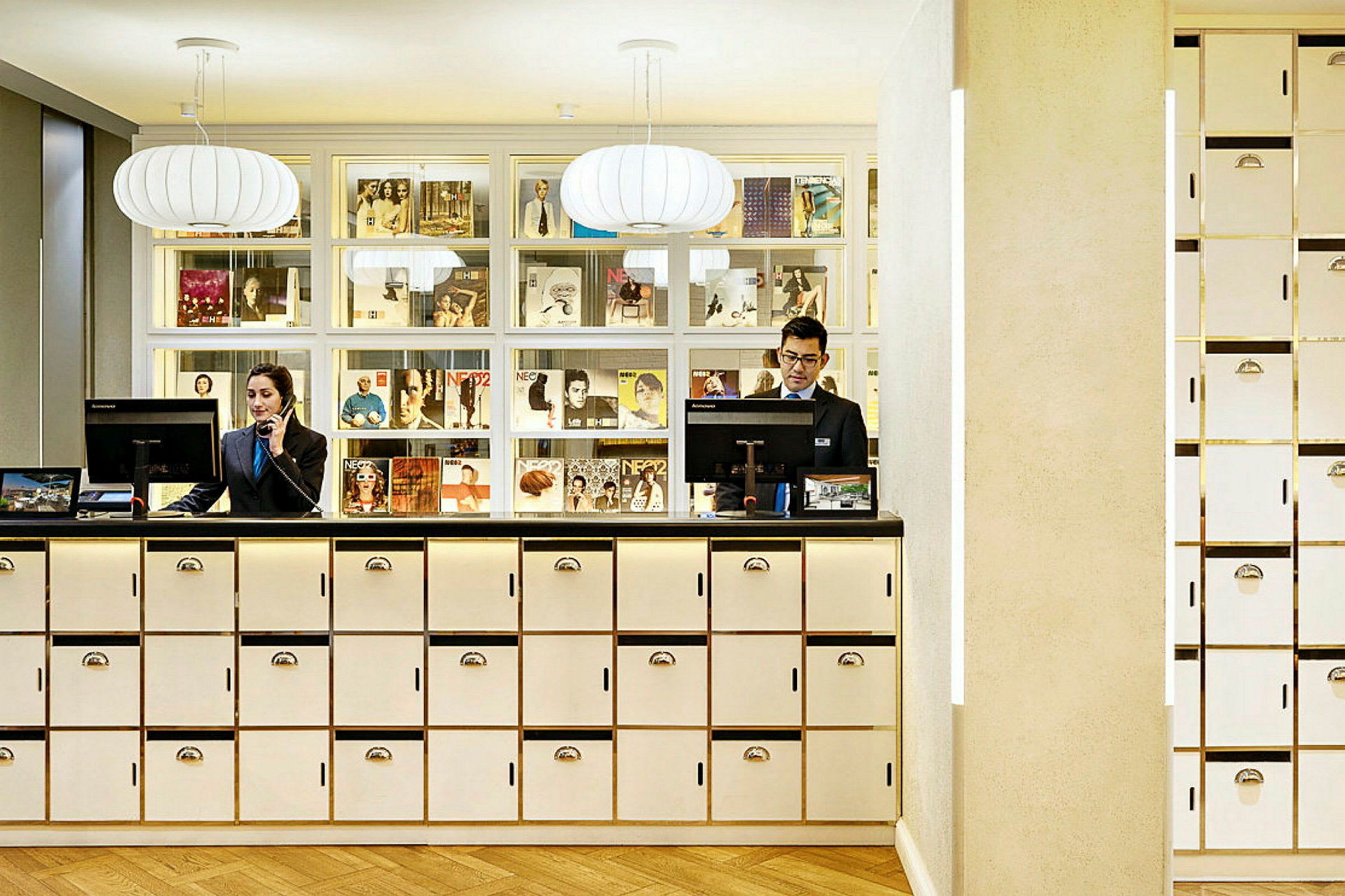 Reception med hvide skabe og dekorativ belysning, bag receptionsskranken er der en væghylde med blade og magasiner. (AI-genereret tekst)