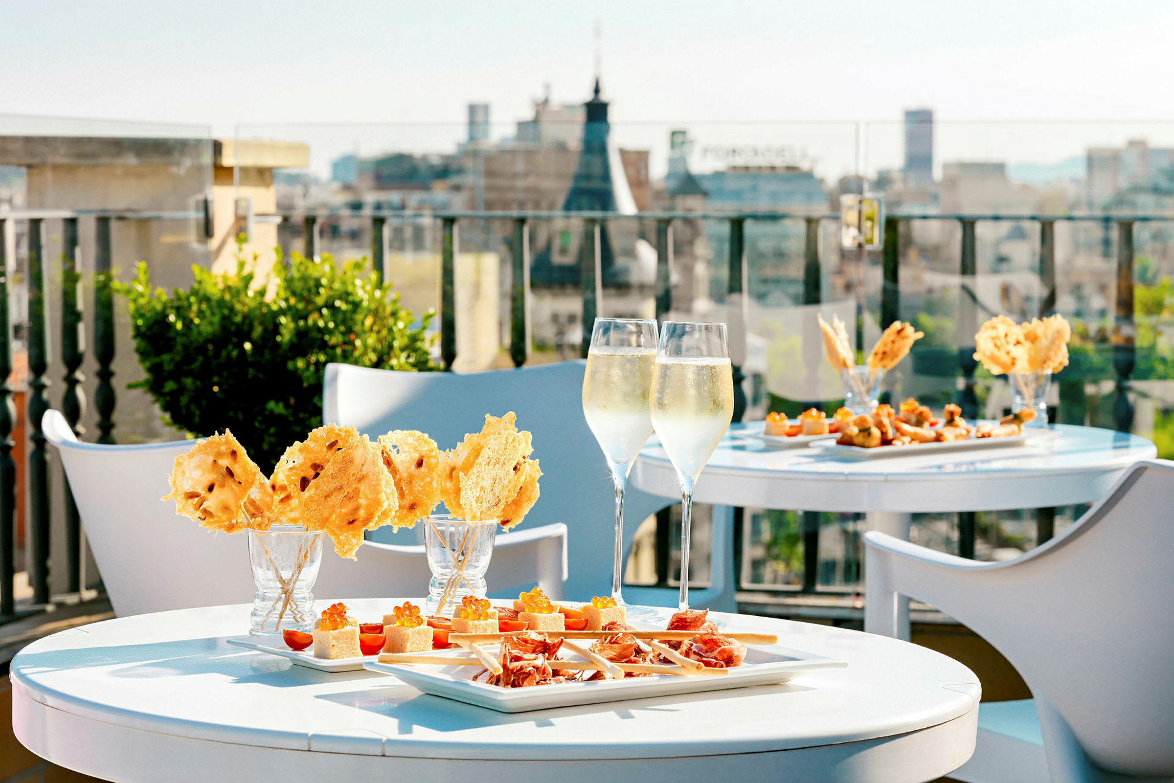En udendørs terrasse med borde serveret med snacks og to glas kølig drik, med en udsigt over byen i baggrunden. (AI-genereret tekst)