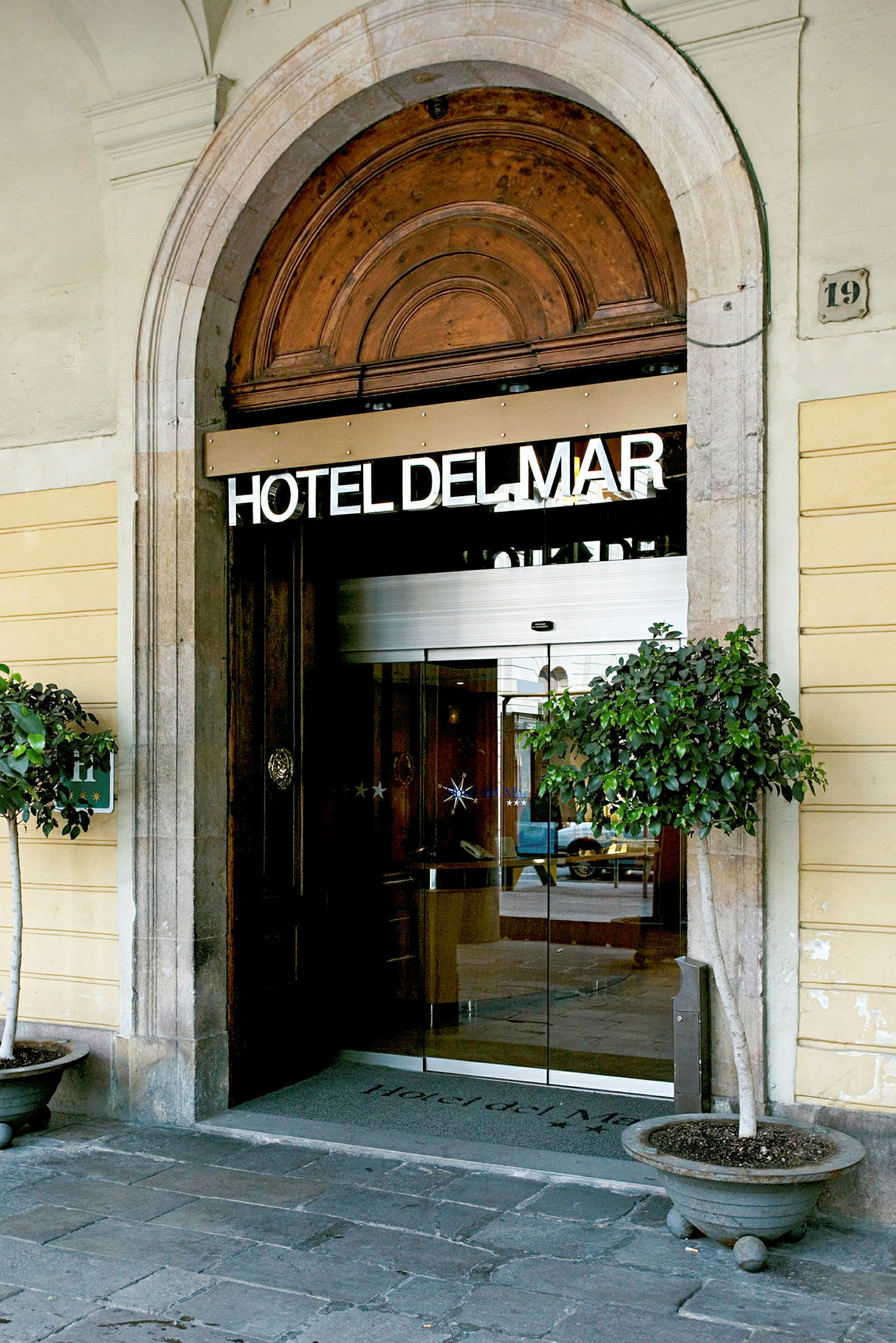 En indgang til et hotel markeret med teksten 'HOTEL DEL MAR'. Indgangen er hvælvet med glasdøre og har dekorative blomsterkrukker på hver side. (AI-genereret tekst)