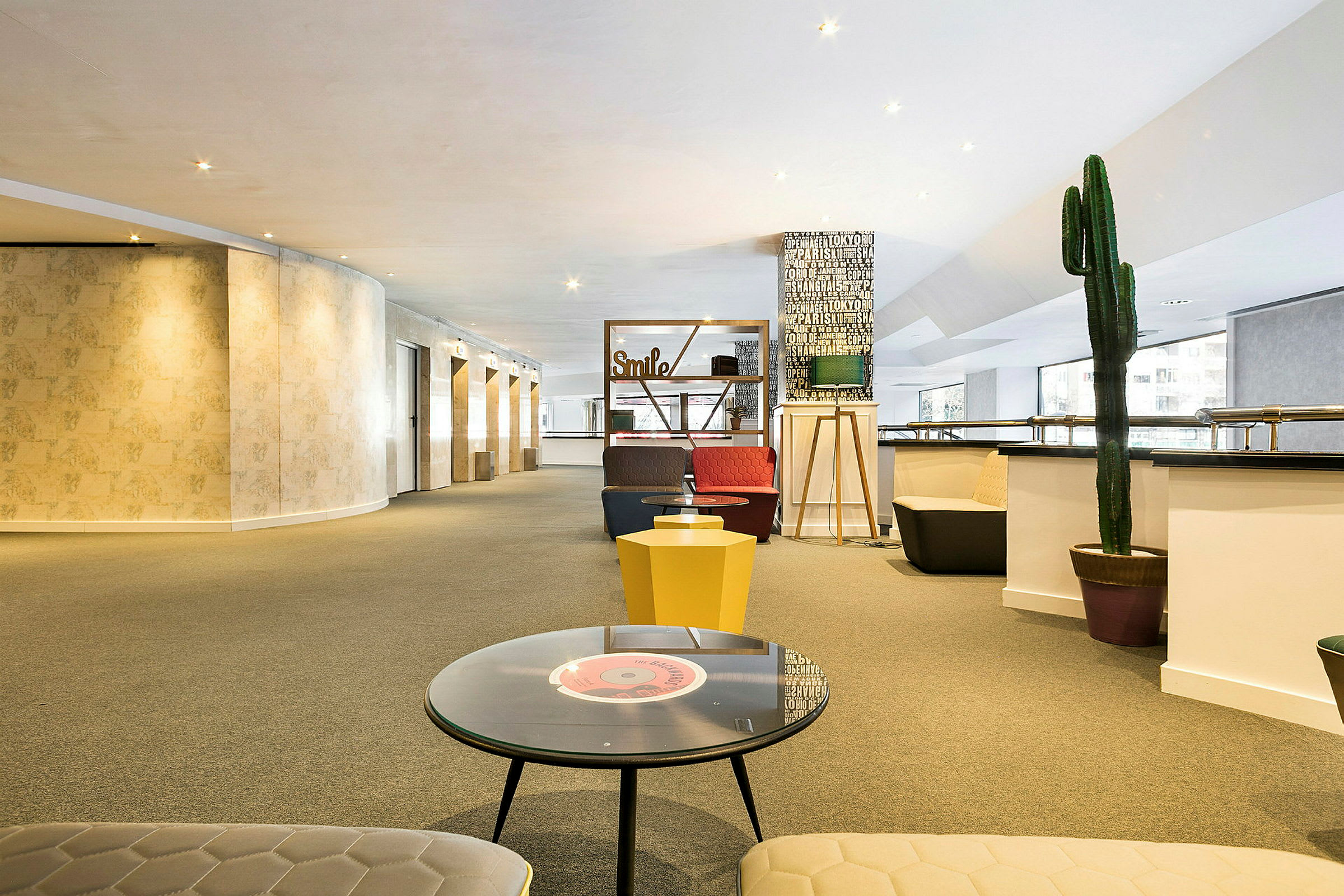 En rummelig og lys lounge med siddepladser, borde og dekorationer, herunder en stor kaktus og skilte med tekst. (AI-genereret tekst)
