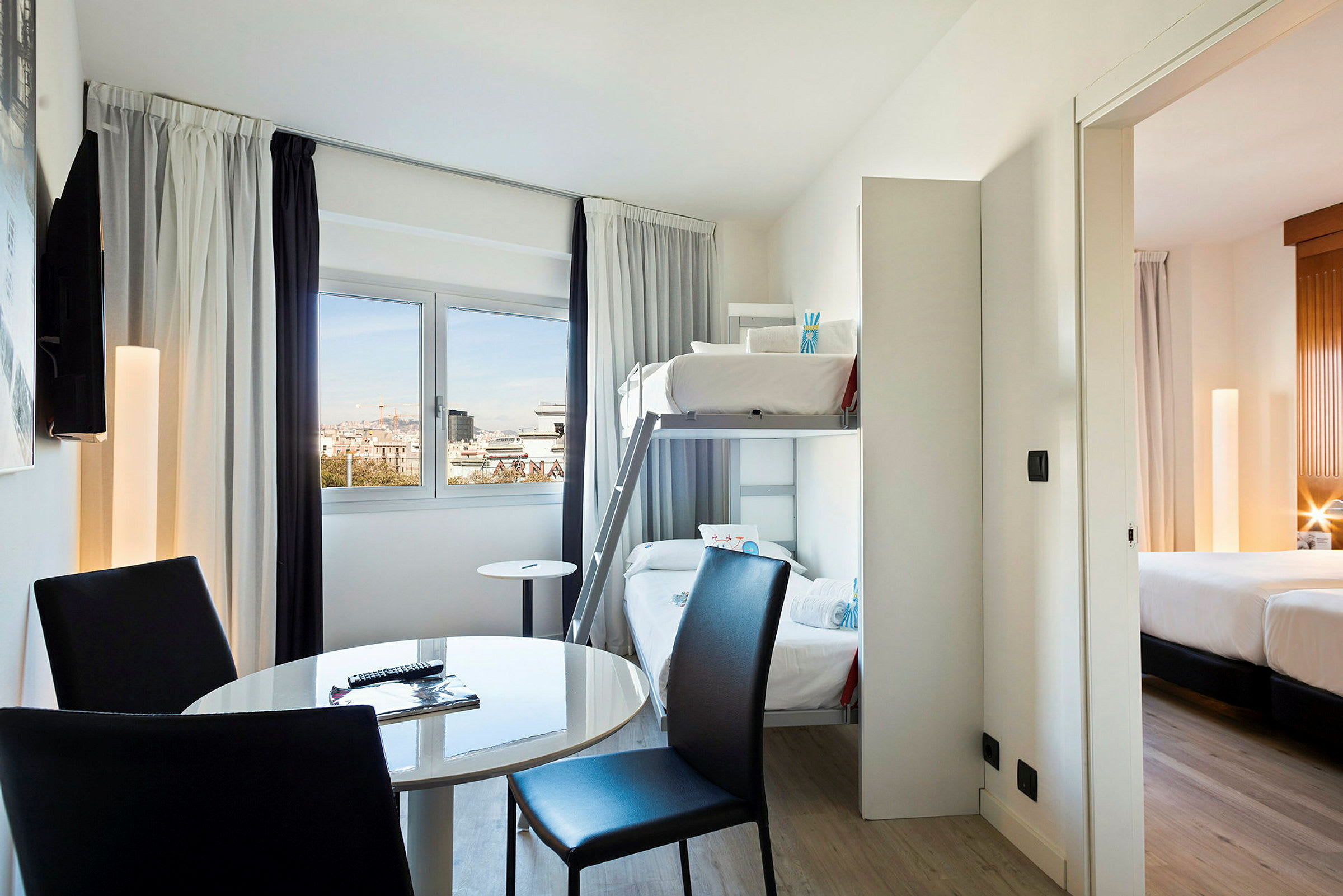 Et hotelværelse med køjesenge, et rundt bord og stole samt en udsigt gennem vinduet. Et tilstødende soveværelse med dobbeltseng ses. (AI-genereret tekst)