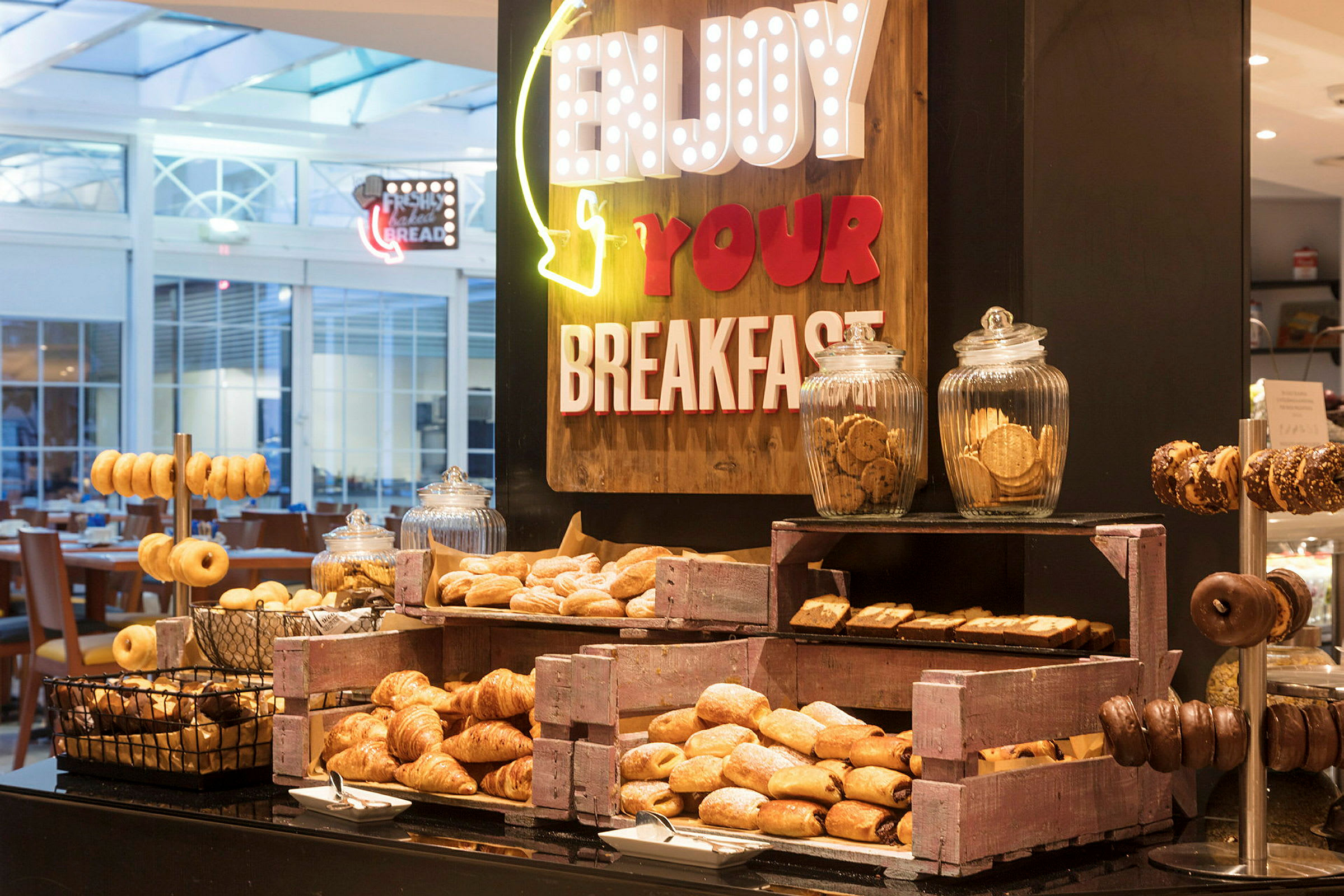 En buffet med forskellige morgenmadsprodukter som croissanter, donuts og småkager, arrangeret med skilte der opfordrer til at nyde morgenmaden. (AI-genereret tekst)