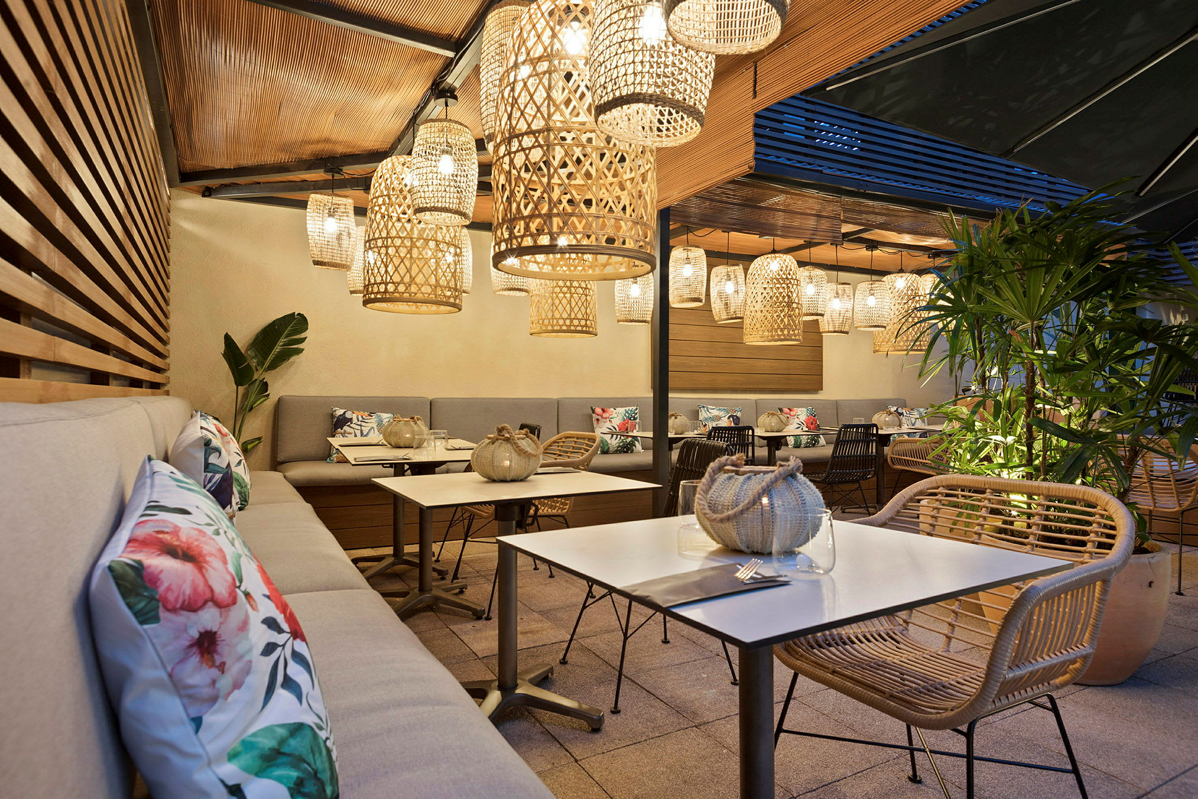 Terrasse med siddepladser, borde og dekorative kurvelamper, der hænger fra loftet. Planter og pyntepuder er synlige. (AI-genereret tekst)