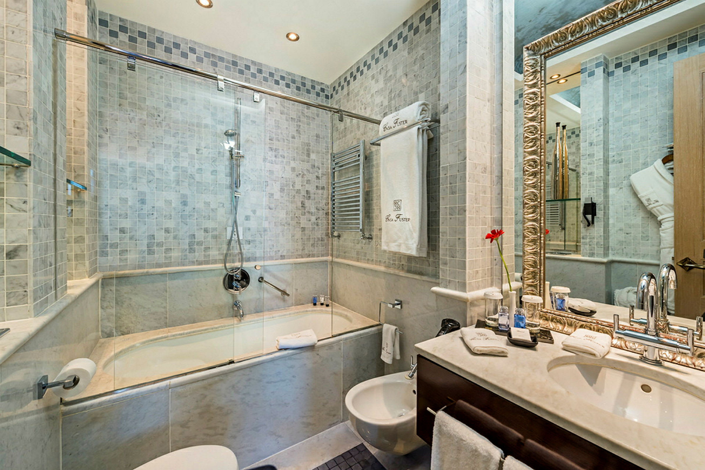 Et badeværelse med en brusekabine kombineret med badekar, en vask med et stort spejl og en bidet. Væggene er flisebelagte og der er håndklæder og andre badeværelsesartikler. (AI-genereret tekst)