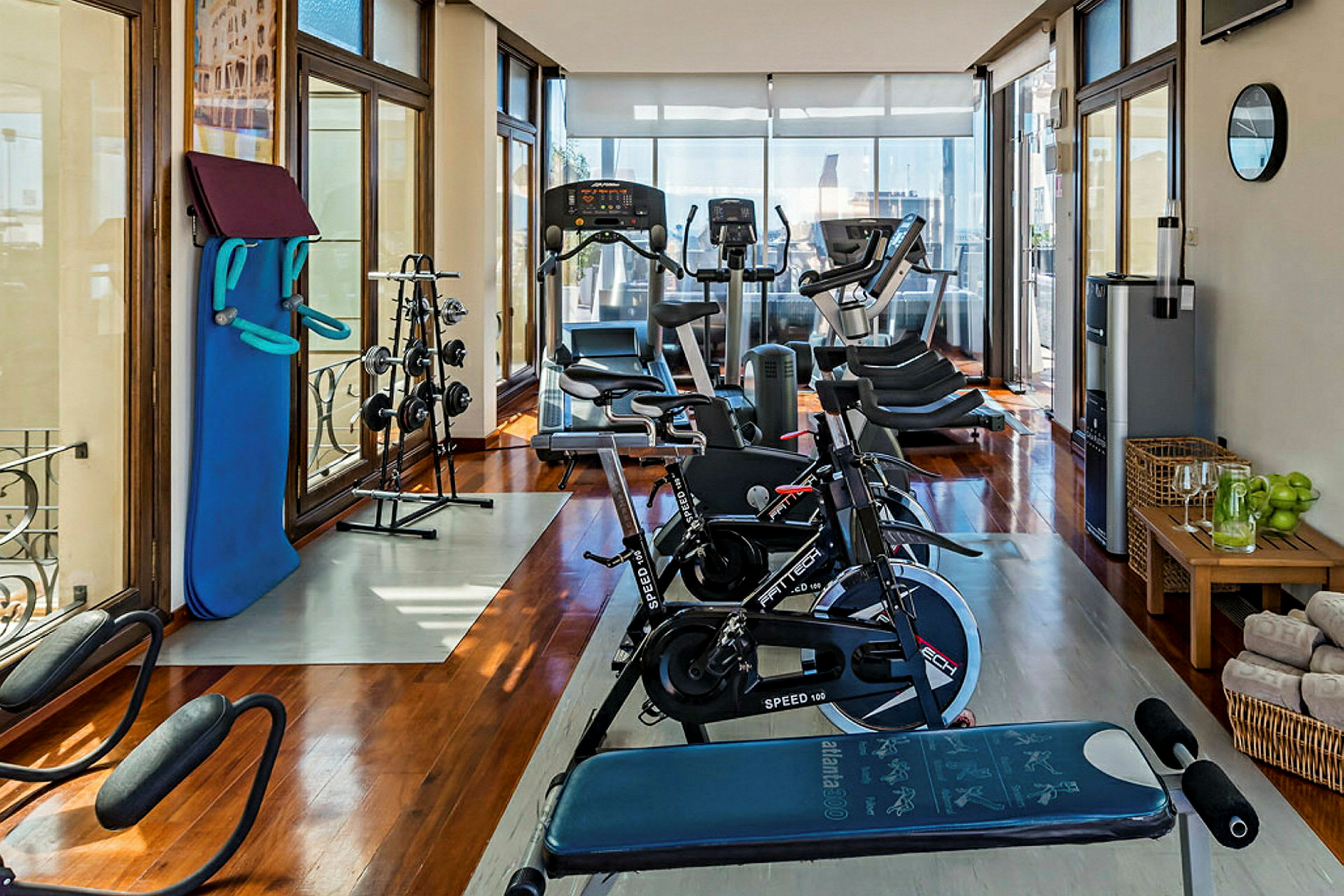 Et lyst fitnesscenter med træningsudstyr, herunder motionscykler, løbebånd, vægte og en bænk samt store vinduer og komfortable faciliteter. (AI-genereret tekst)