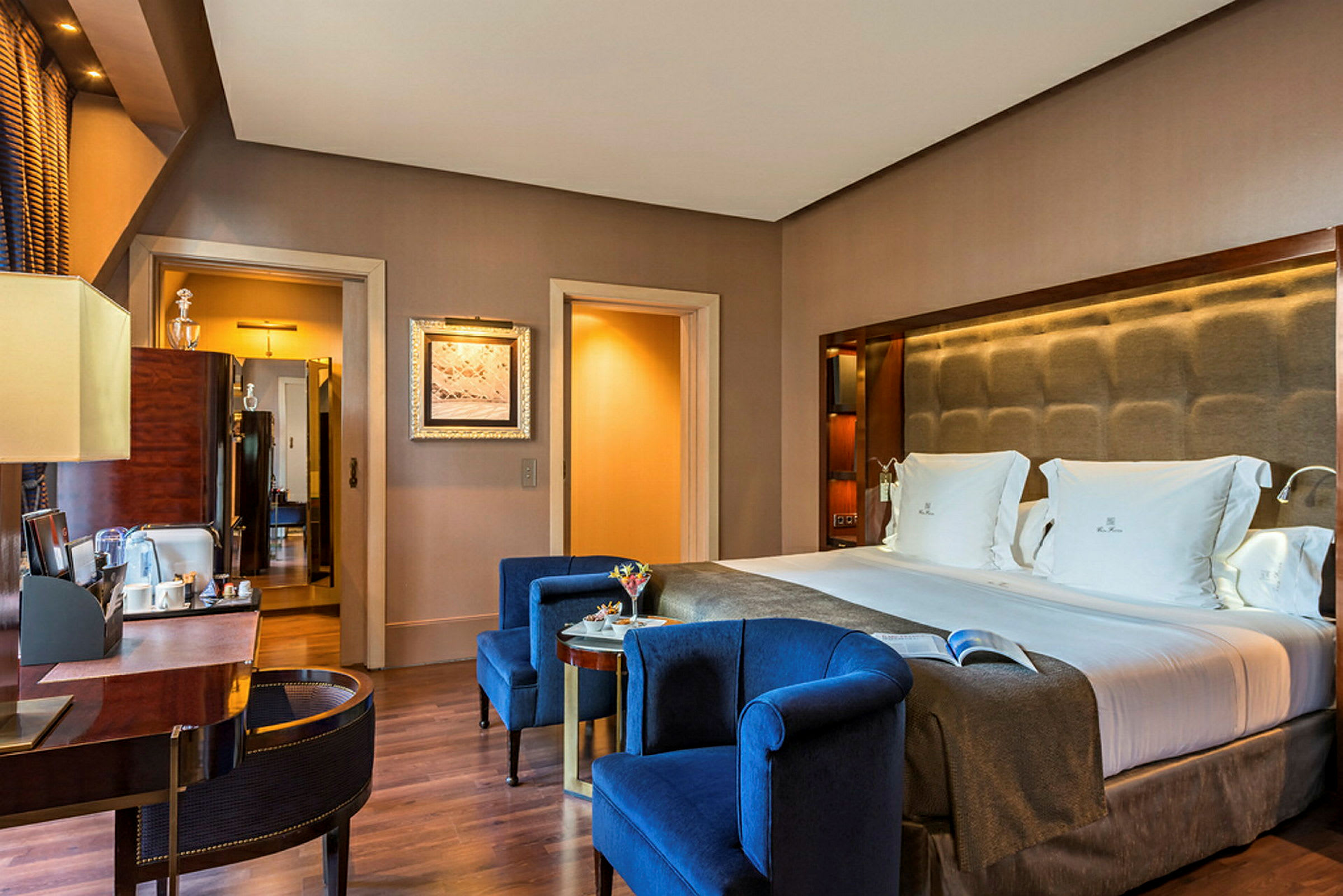 Et elegant hotelværelse med en dobbeltseng og blå lænestole foran et bord. (AI-genereret tekst)