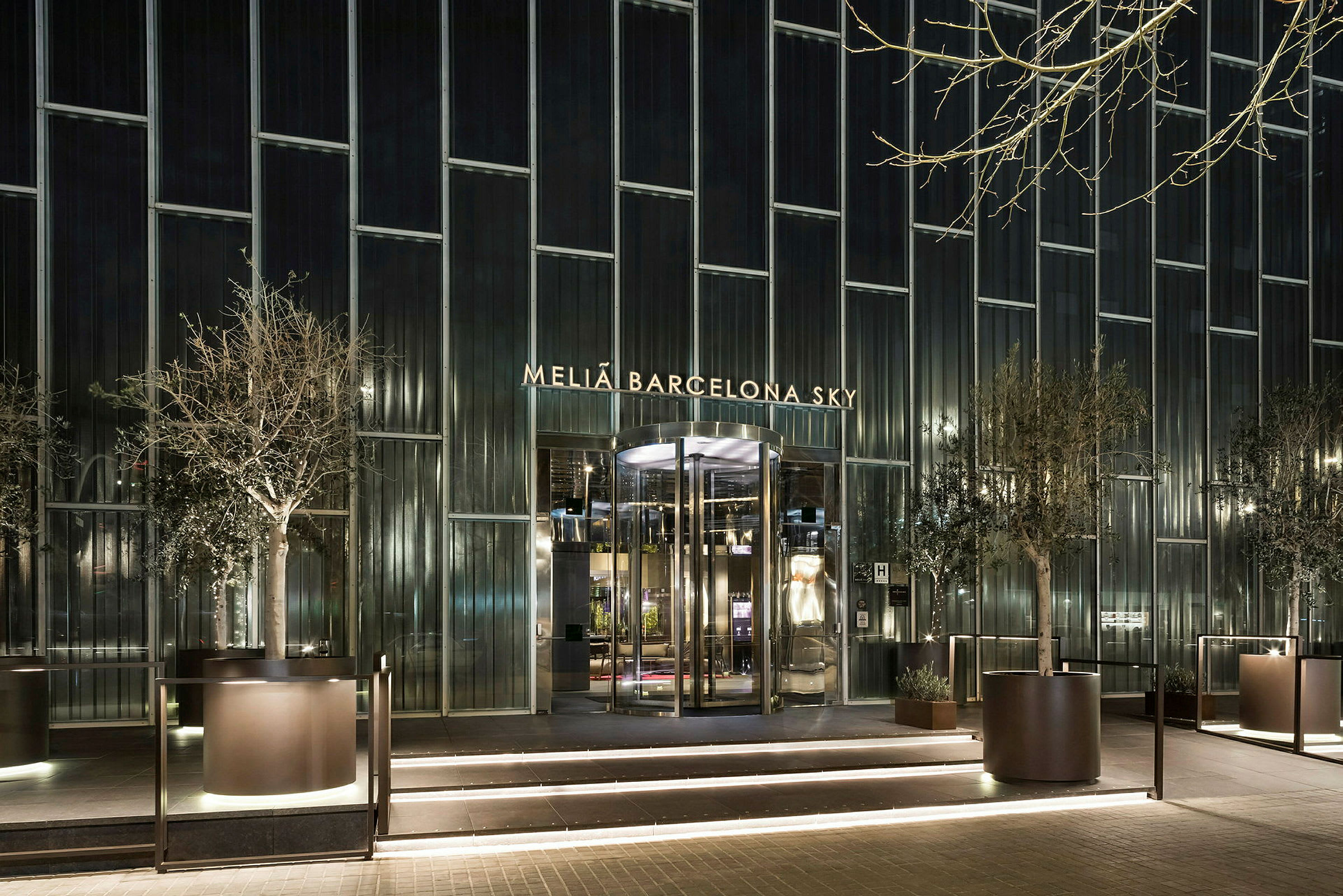 Facaden af Melia Barcelona Sky Hotel med oplyst indgang og store glasvinduer, omgivet af plantede træer. (AI-genereret tekst)