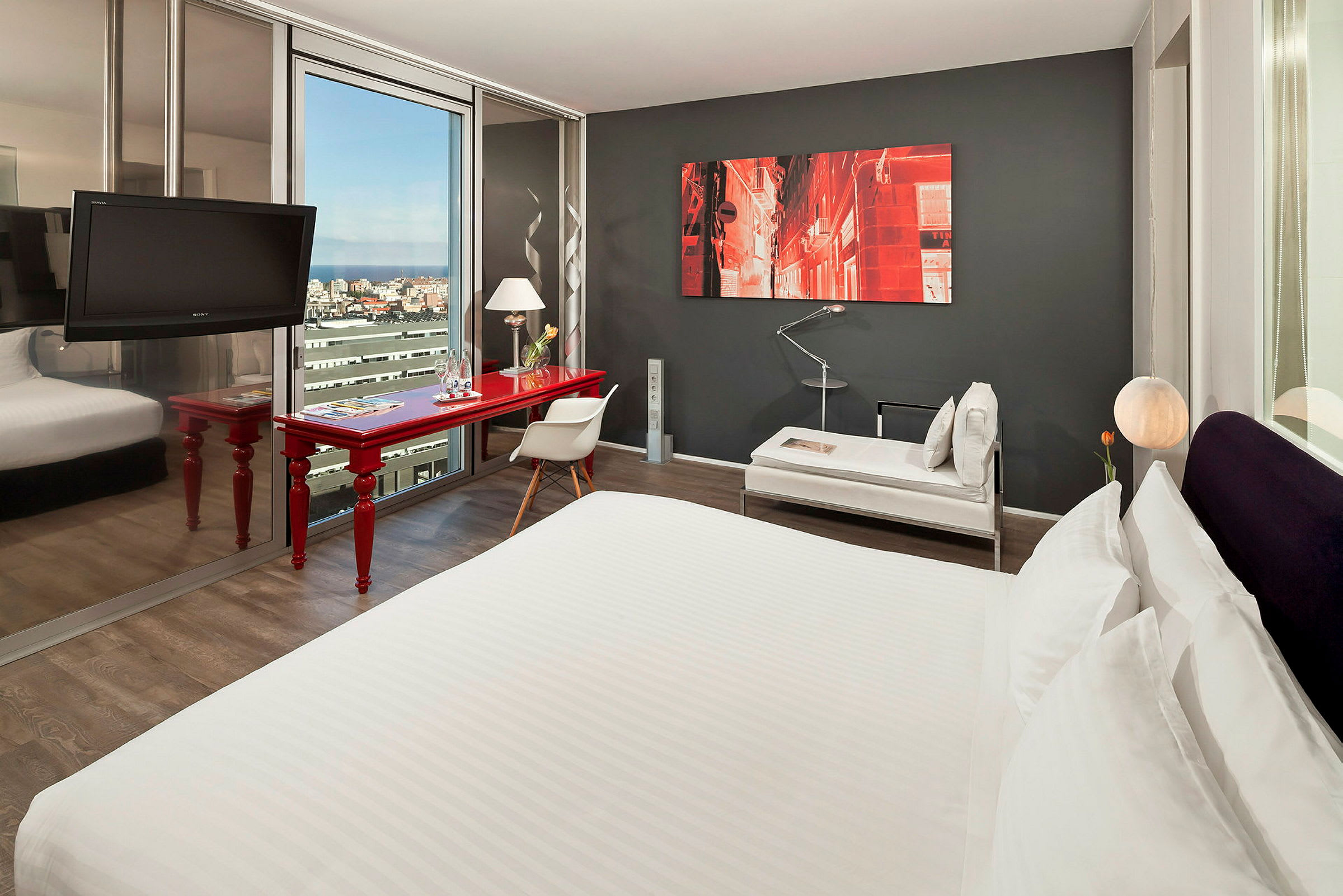 Et moderne hotelværelse med en stor seng, et rødt skrivebord, en stol og et sofabord. Et maleri pryder den mørkegrå væg, og et stort vindue viser udsigt over byen. (AI-genereret tekst)