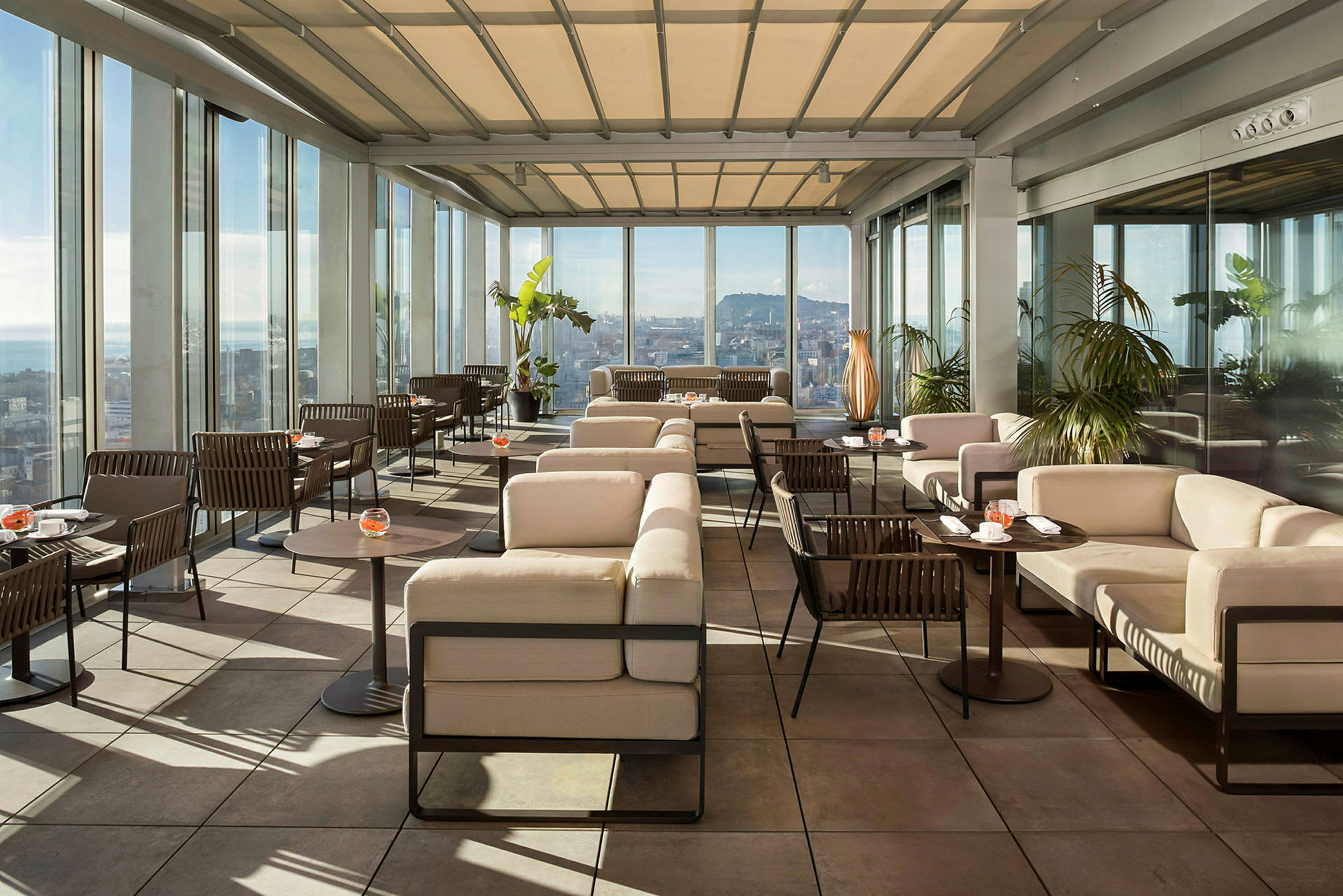En moderne tagterrasse med stilfulde siddepladser og glasvægge med udsigt over en by. Solskinsvejr og grønne planter skaber en behagelig atmosfære. (AI-genereret tekst)