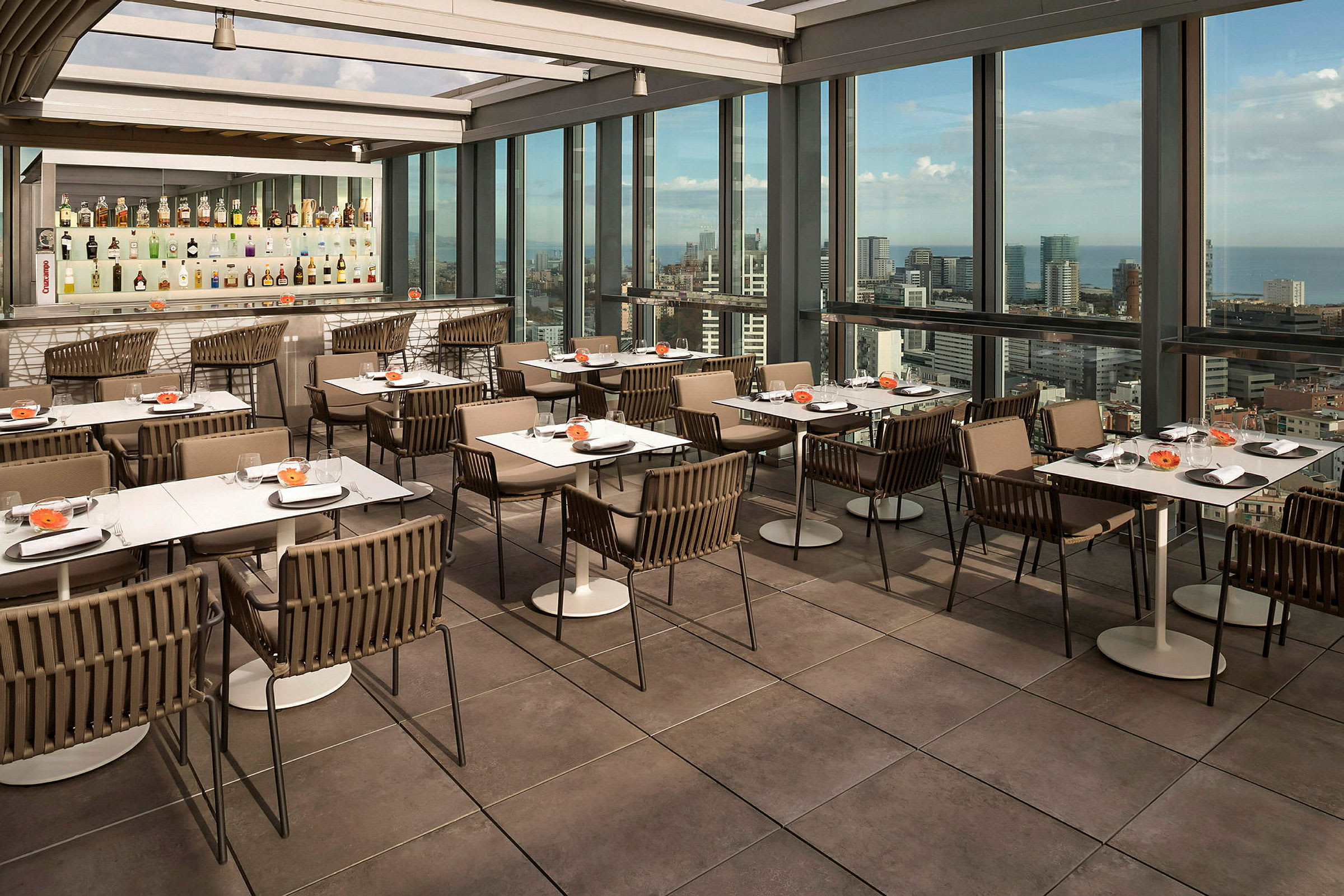 En restaurant med borde og stole på en terrasse med store glasvinduer, der viser en byudsigt. (AI-genereret tekst)