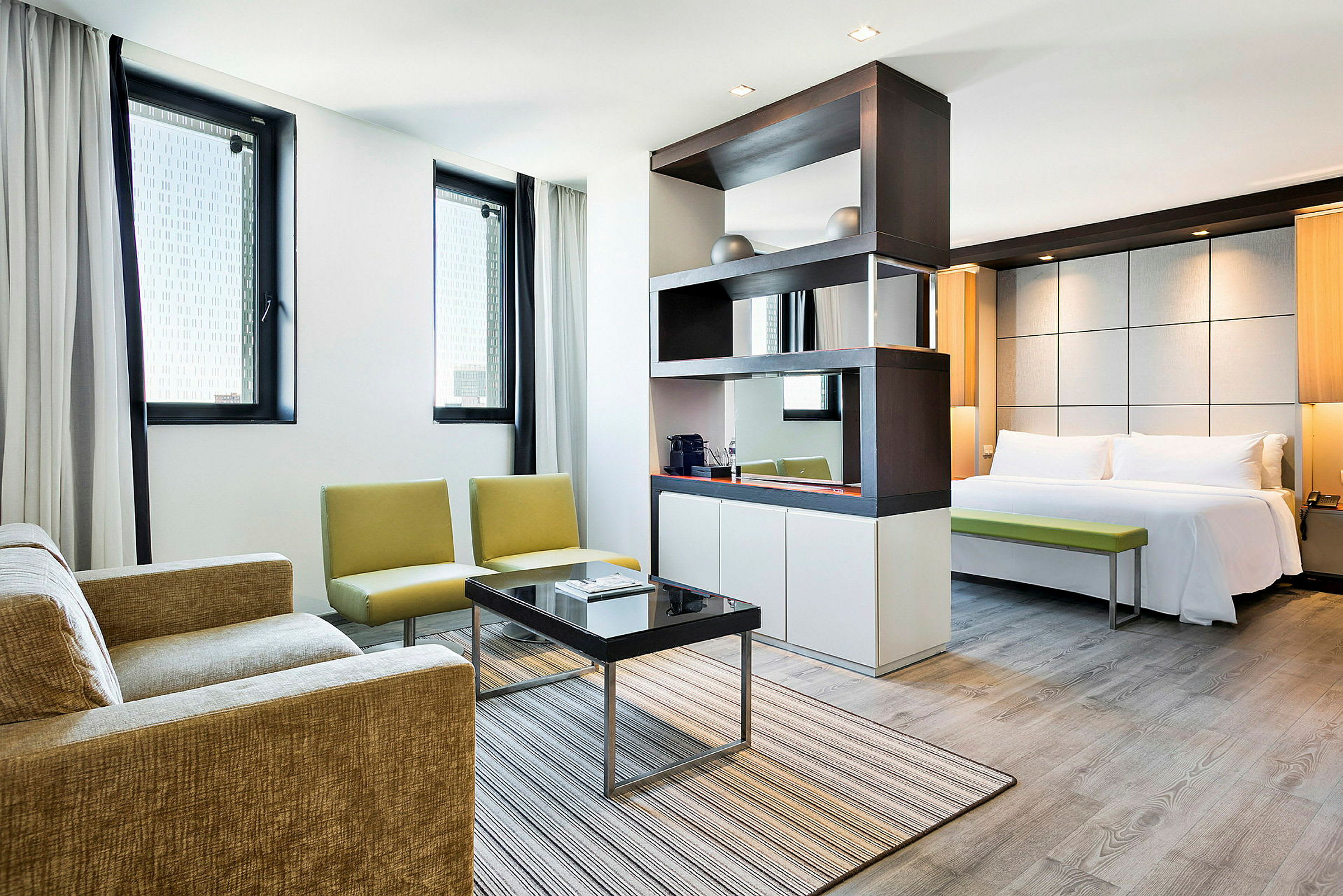 Et moderne hotelværelse med en stue og et soveværelse. Stuen har en sofa, lænestole og sofabord. Soveværelset har en dobbeltseng og en bænk. (AI-genereret tekst)