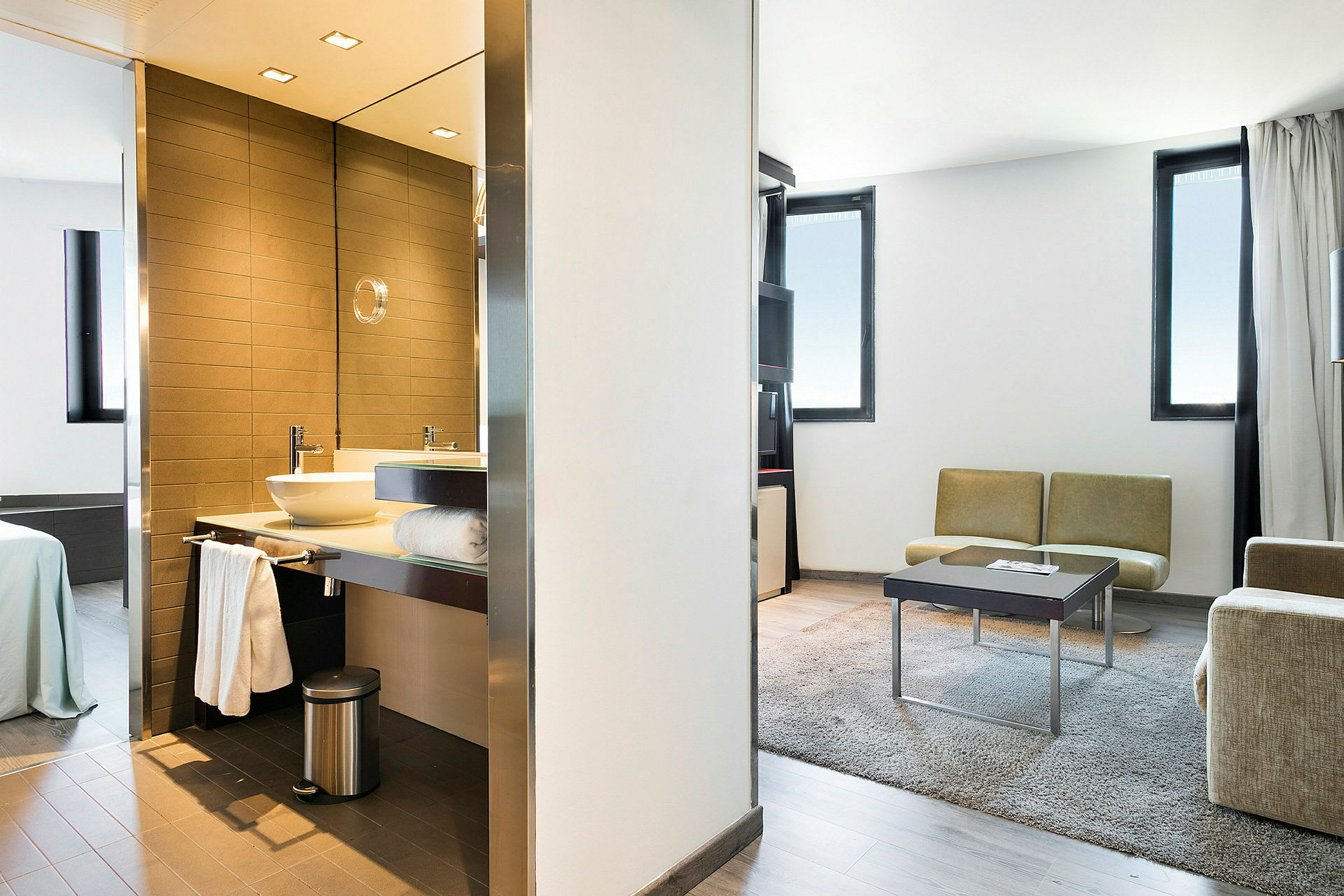 Et moderne hotelværelse med badeværelse med håndvask og spejl til venstre, soveværelset med seng længere bagved, samt stuen med sofaer, bord og tæppe til højre. (AI-genereret tekst)