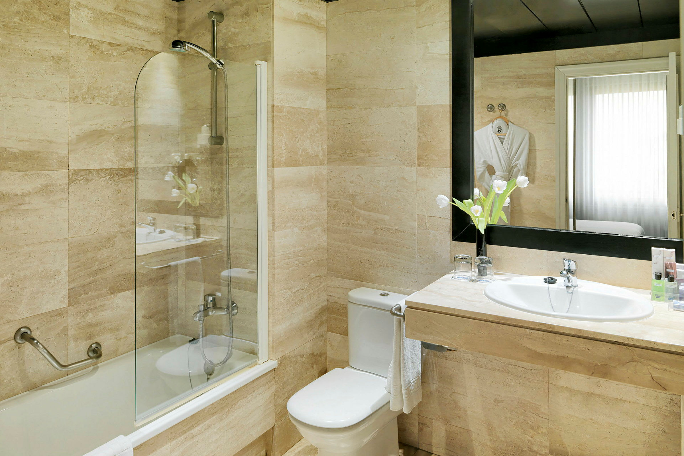Badeværelse med badekar, bruser bag glasvæg, toilet, håndvask og spejl ovenover. Blomster på håndvasken. (AI-genereret tekst)