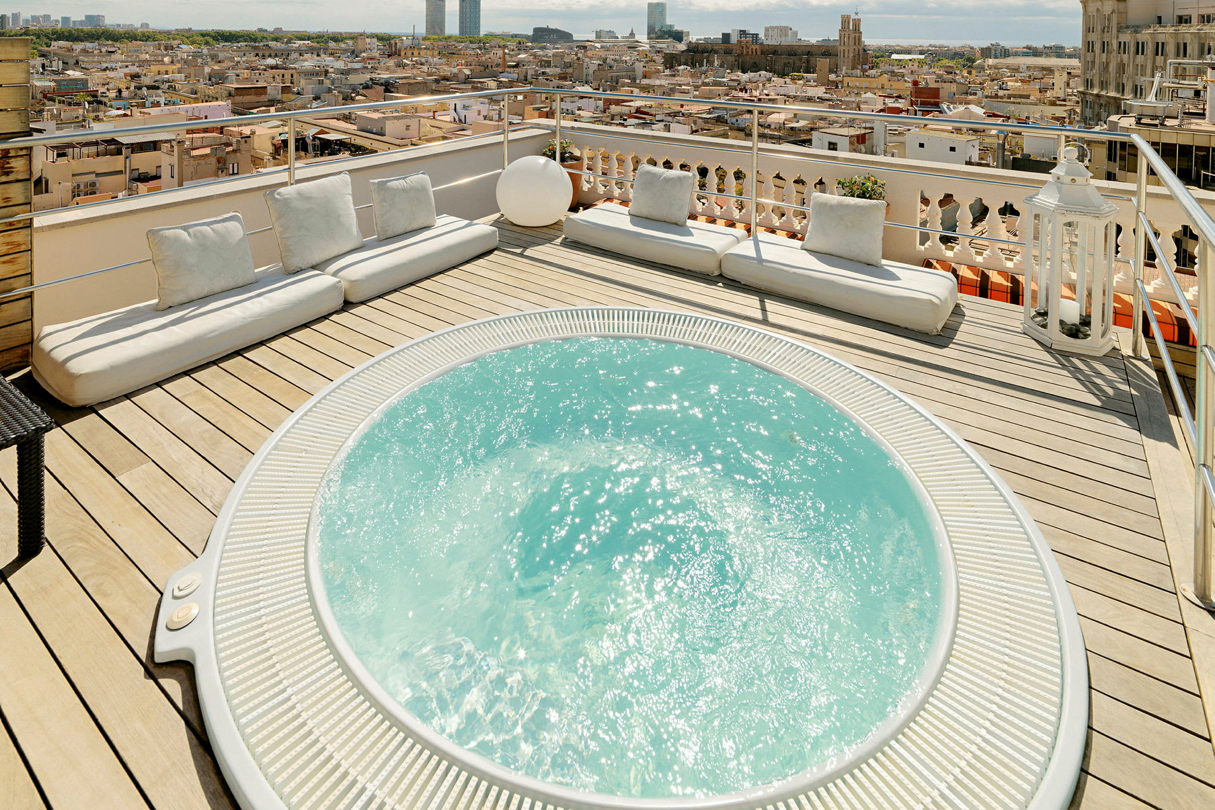 En terrasse med en jacuzzi og flere hvide siddepuder, med udsigt over en by. (AI-genereret tekst)