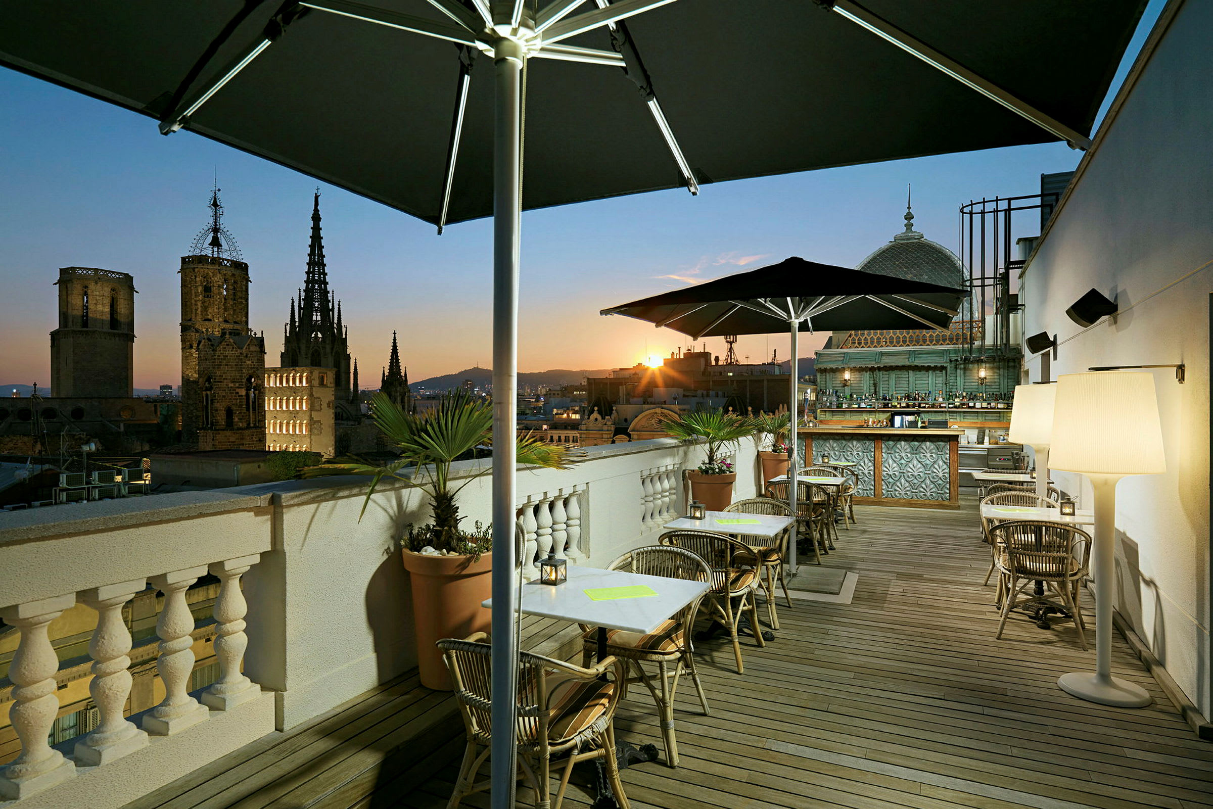 En tagterrasse med borde og stole, parasoller og planter, med udsigt over en by med katedral i solnedgangen. (AI-genereret tekst)