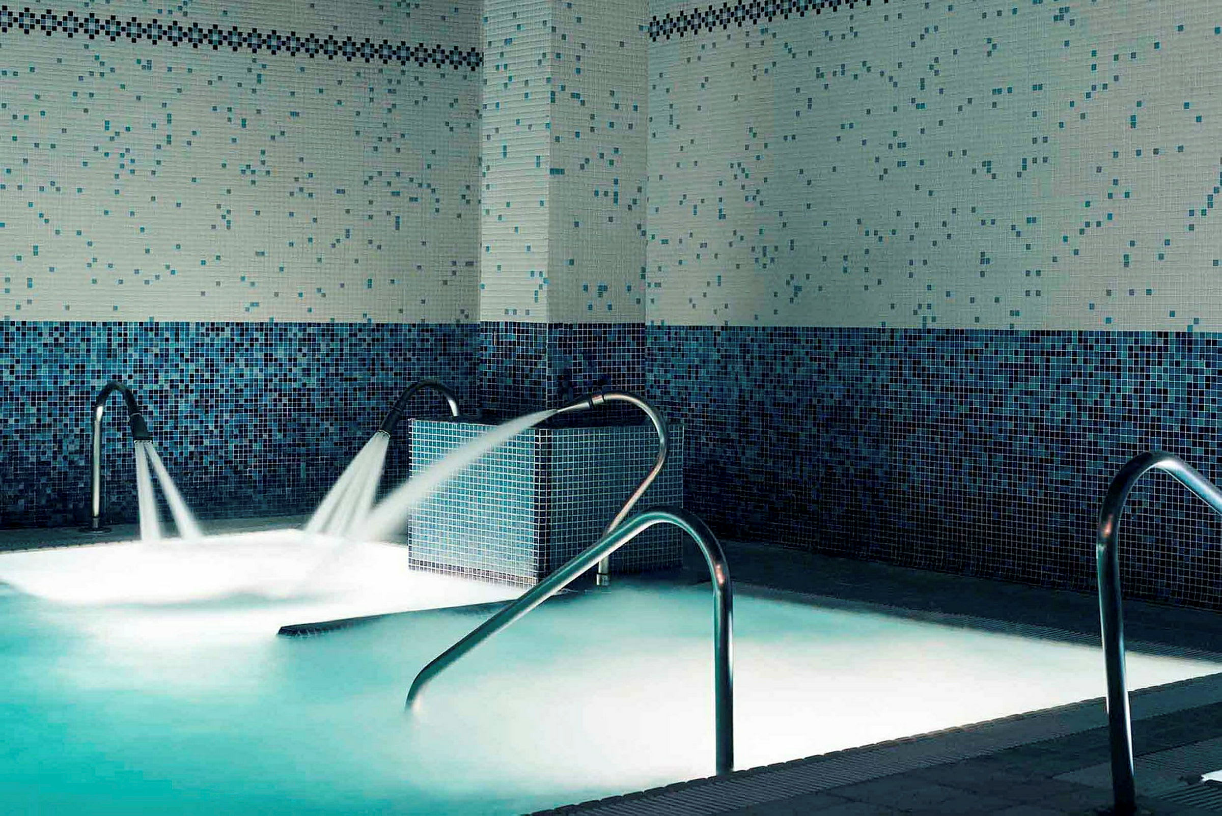 Indendørs pool med blå mosaikvægge og metalgelændere, vand sprøjter ud fra flere udløb. (AI-genereret tekst)
