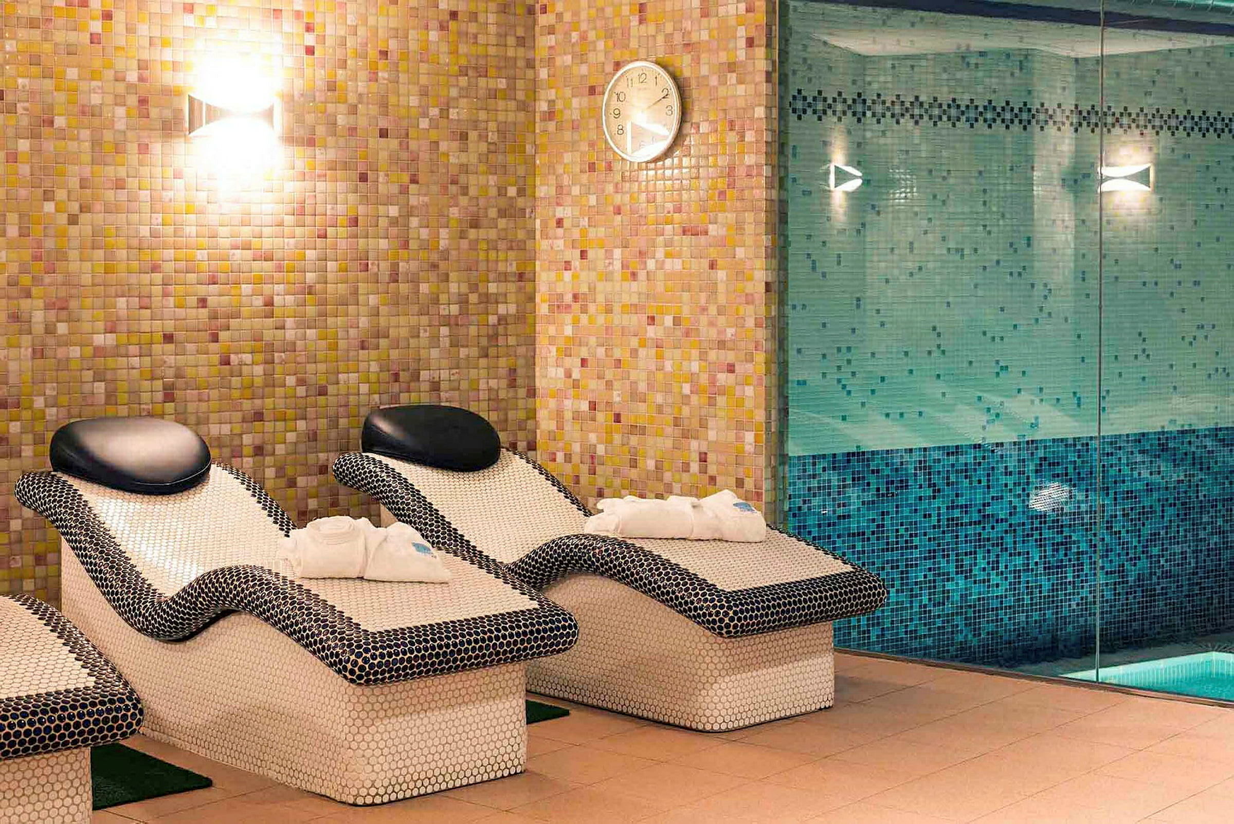 Billede af et afslapningsområde med mosaikbeklædte liggestole og en pool bag glasvæg. (AI-genereret tekst)