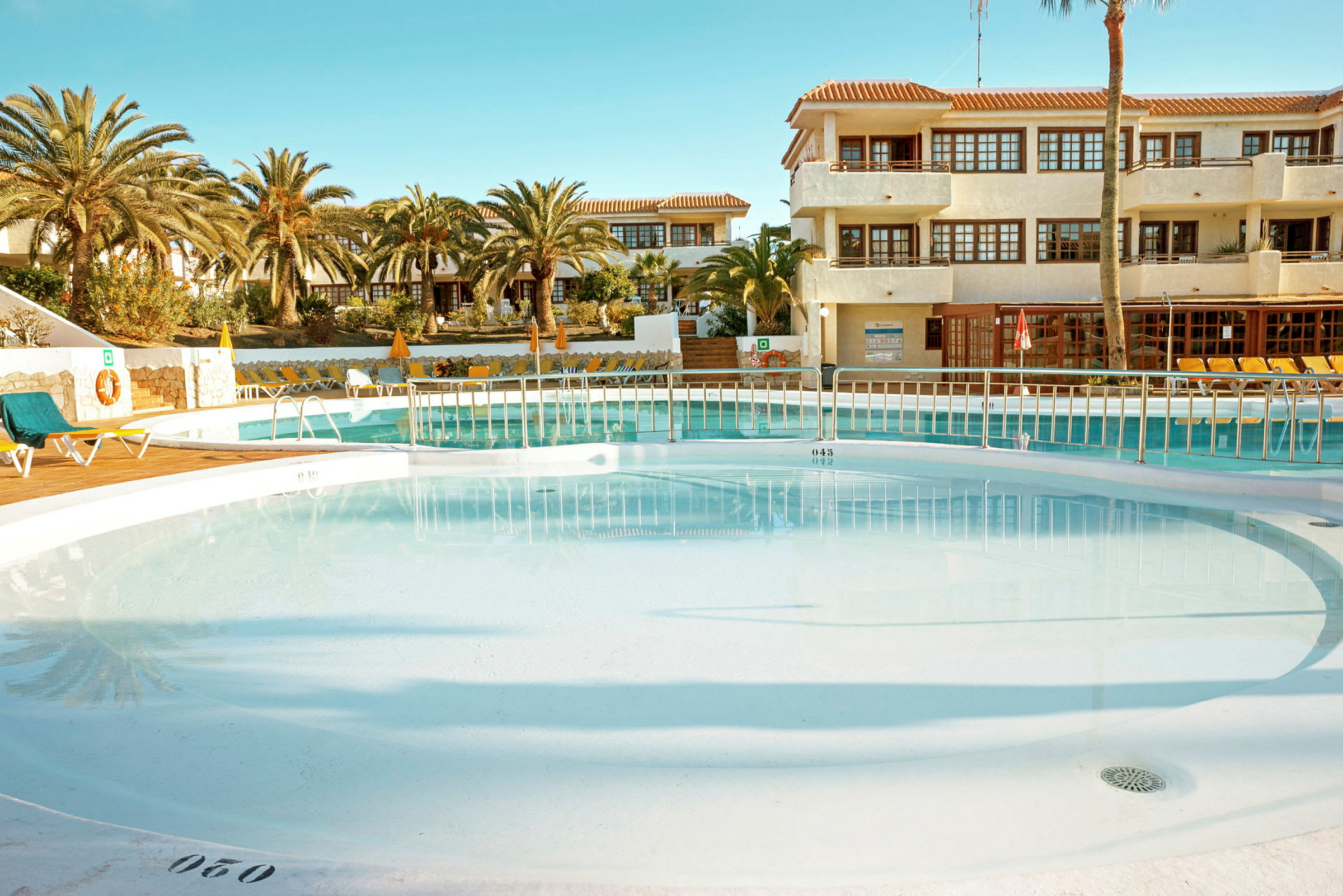 En udendørs pool med solterrasse, liggestole og omgivet af palmer samt bygninger i baggrunden. (AI-genereret tekst)