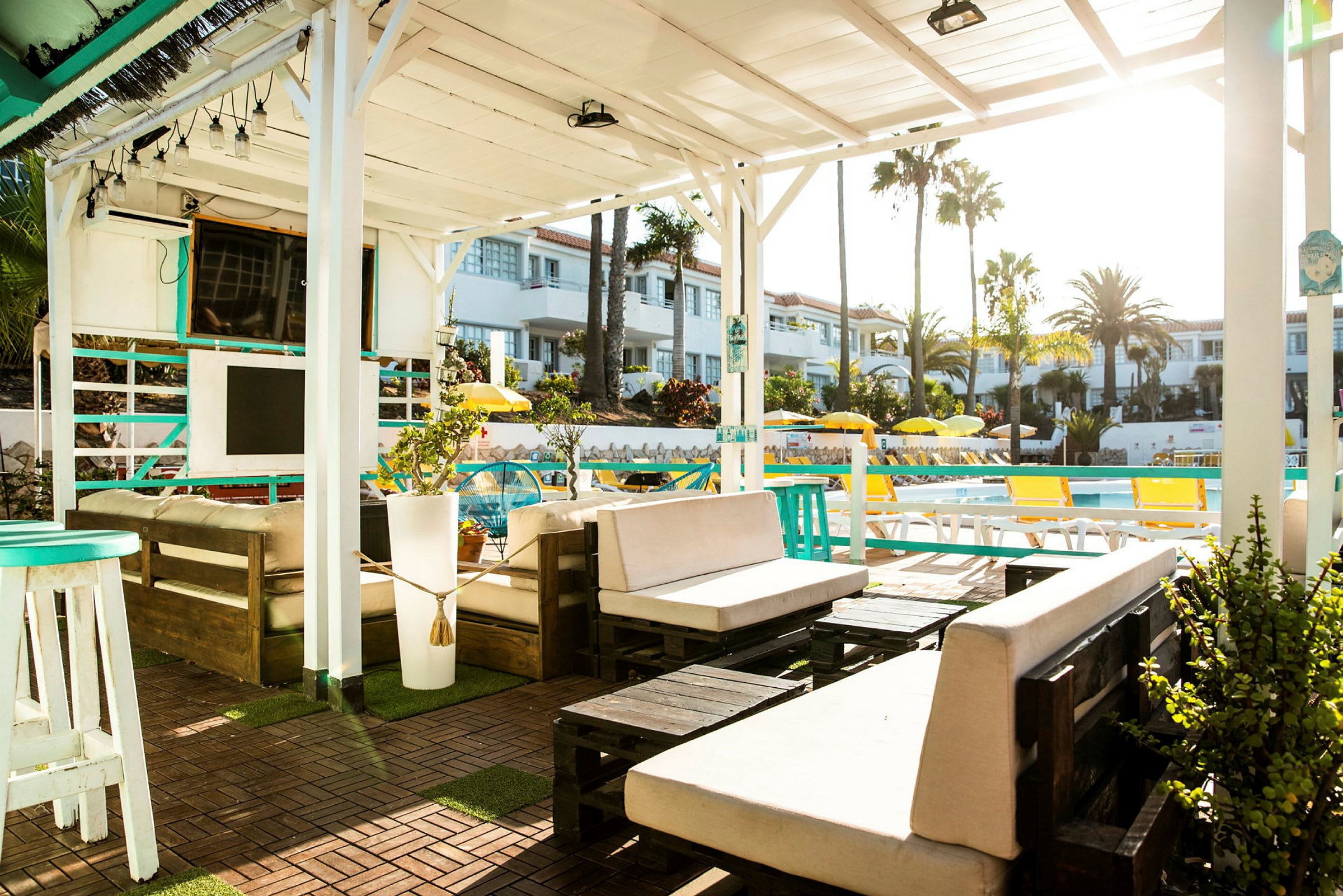 En udendørs lounge under en hvid pergola med møbler og planter, med udsigt over en pool og omkringliggende bygninger. (AI-genereret tekst)