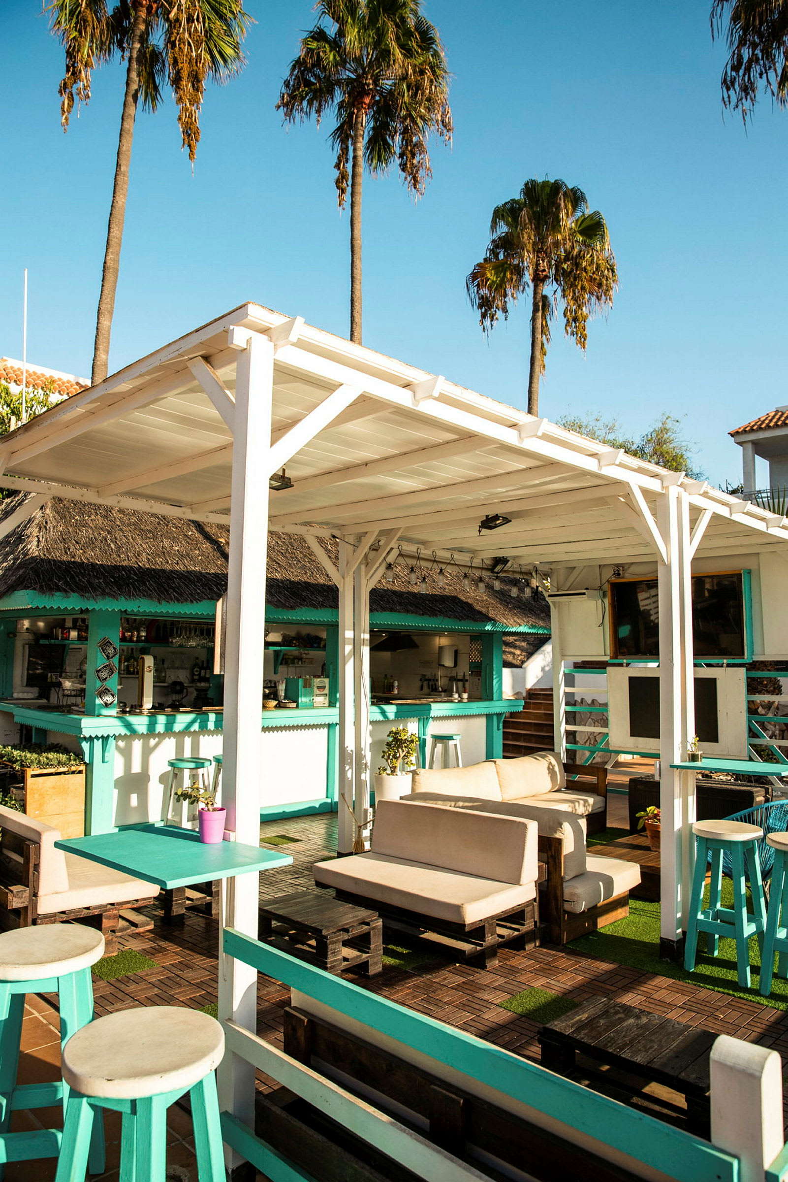 En udendørs lounge med sofaer og borde under en hvid træpergola, omgivet af palmer og en bar. (AI-genereret tekst)