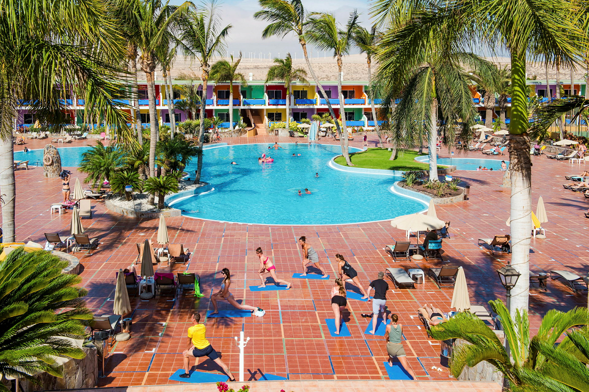 Ved et stort hotel med farverige bygninger dyrker folk udendørs yoga på måtter ved siden af en pool. (AI-genereret tekst)