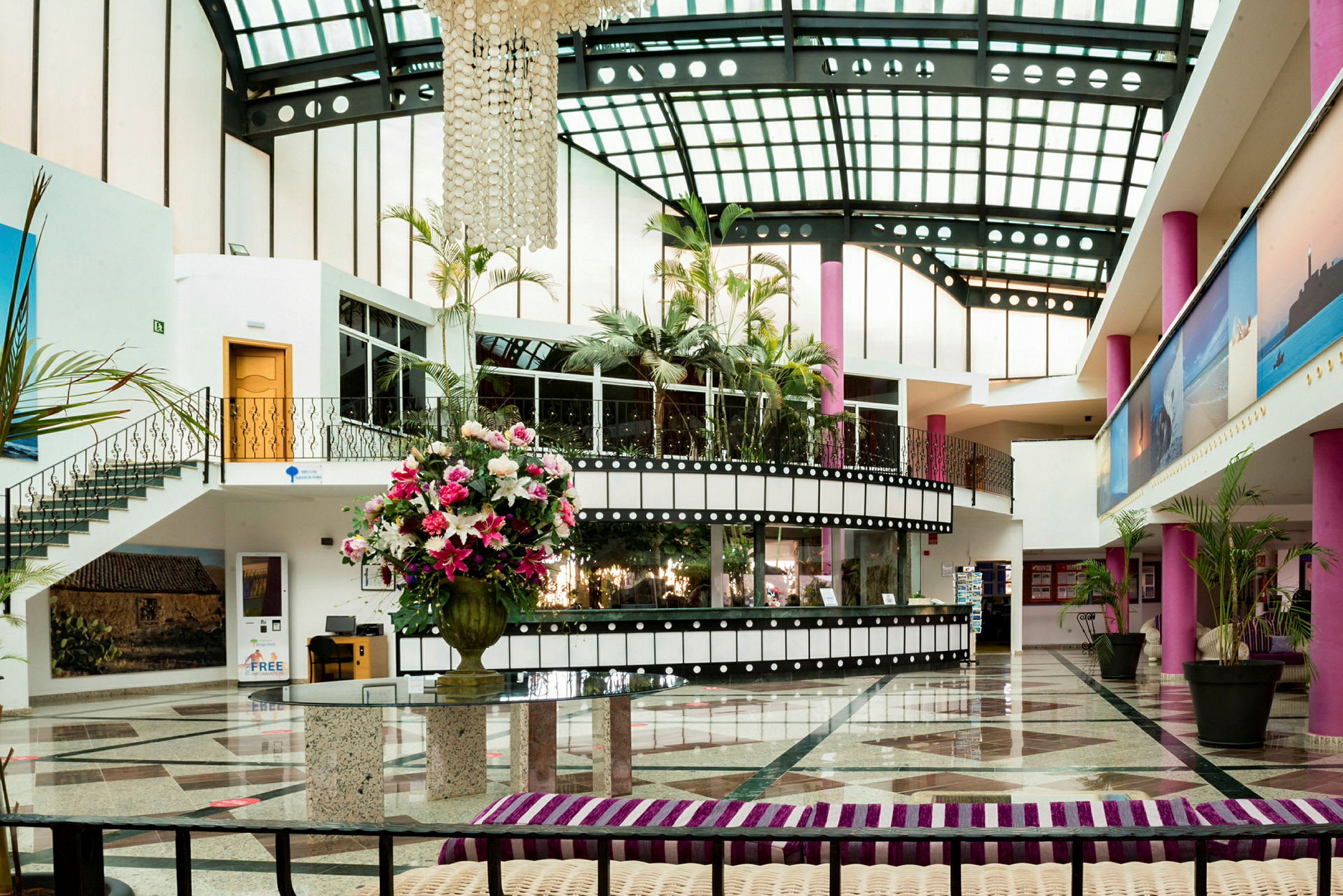 Interiør af en hotellobby med glastag, lyserøde søjler og en stor vase med farverige blomster. (AI-genereret tekst)