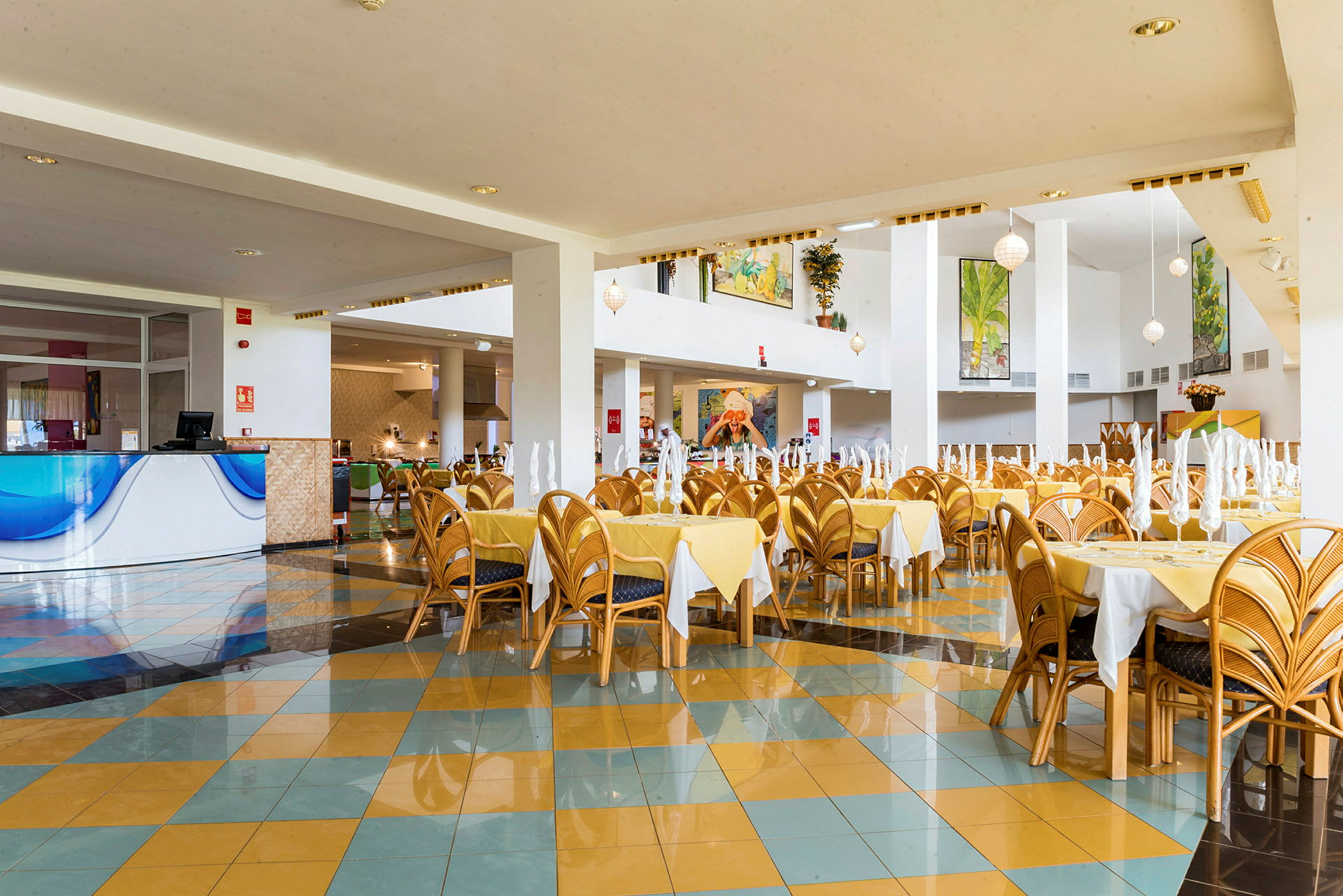 En rummelig restaurant med gule duge og stole af træ. Flere borde er dækket med foldede servietter, og lokalet har fine detaljer og dekorationer. (AI-genereret tekst)
