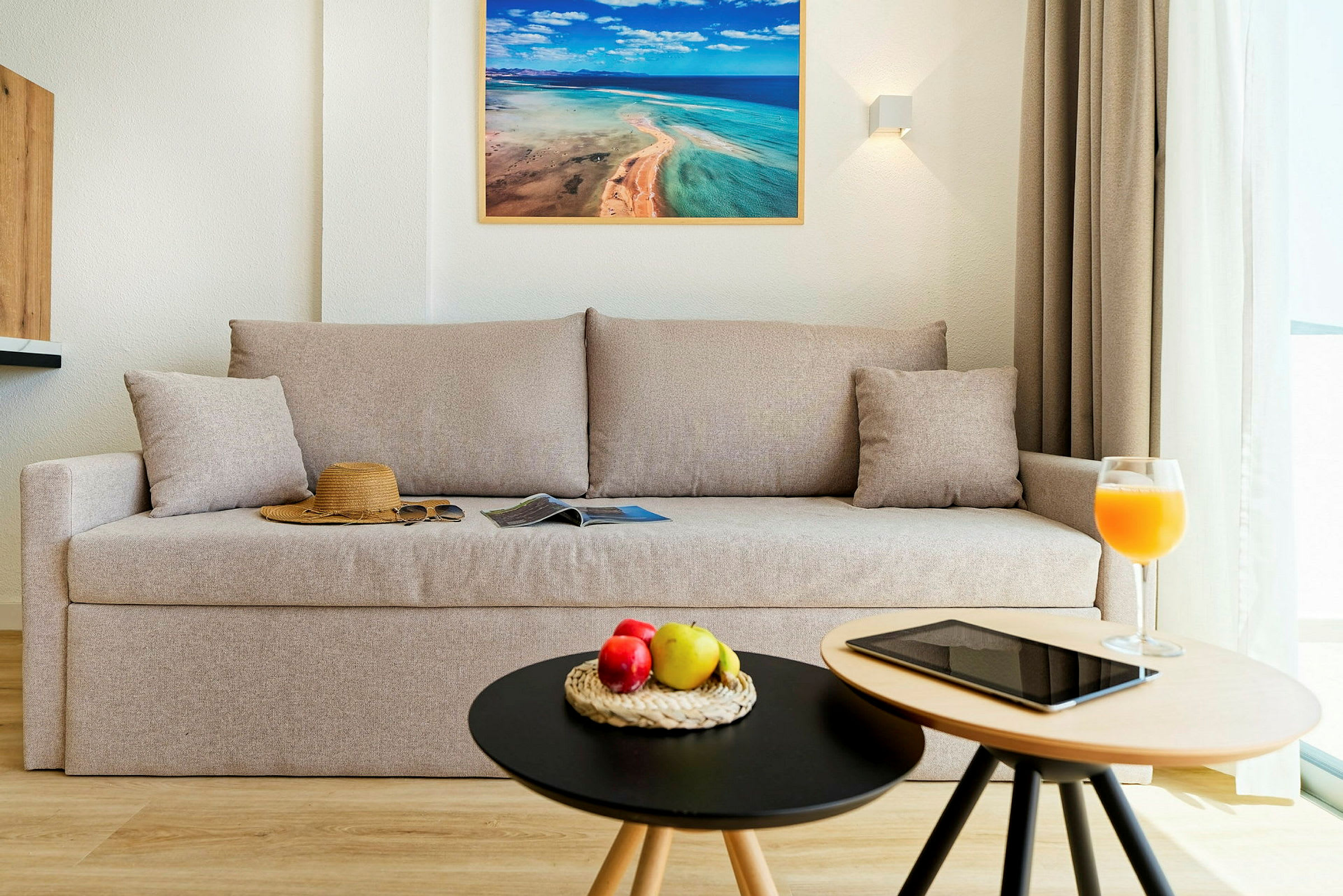 En stue med en beige sofa, en hat og solbriller på sofaen, samt to små borde med frugt, en tablet og et glas juice. (AI-genereret tekst)