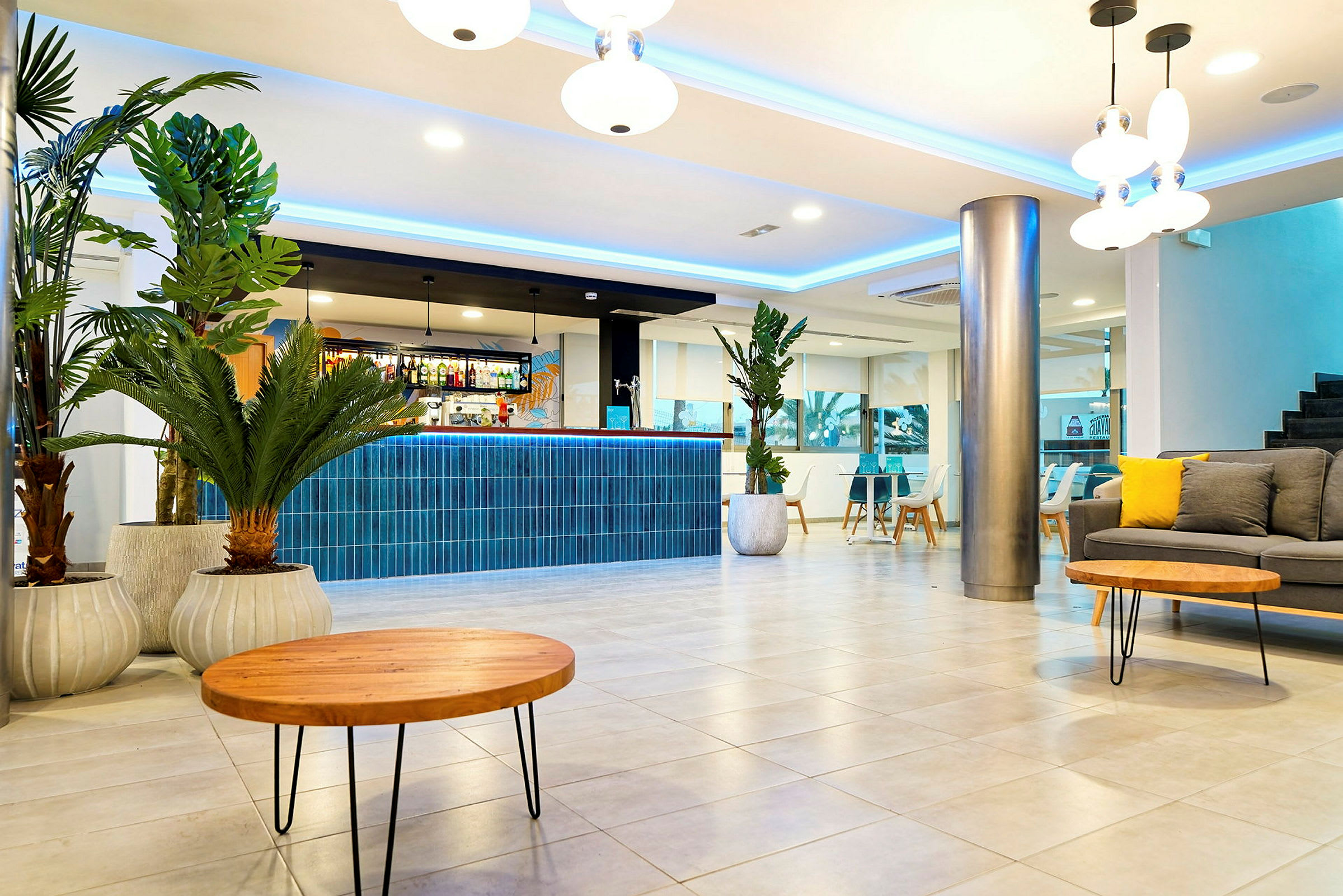 En moderne hotelreception med en skranke dekoreret med blå fliser, nogle grønne planter i krukker, en grå sofa med gule puder og runde træborde. (AI-genereret tekst)