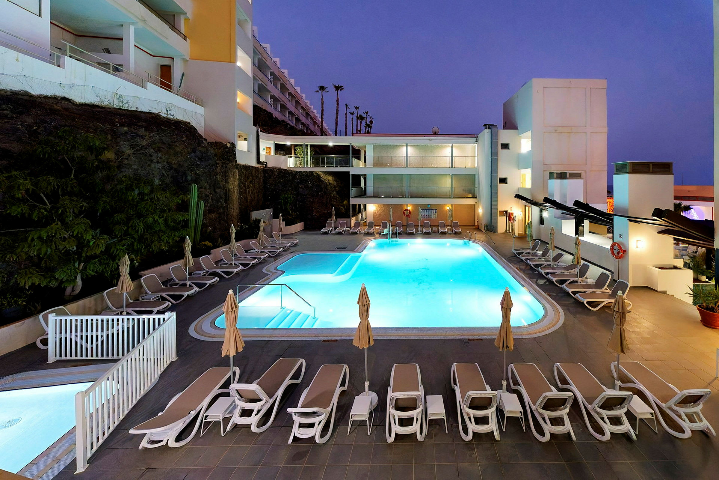 Billedet viser en oplyst hotelpool med omkringliggende solsenge og parasoller om aftenen. (AI-genereret tekst)