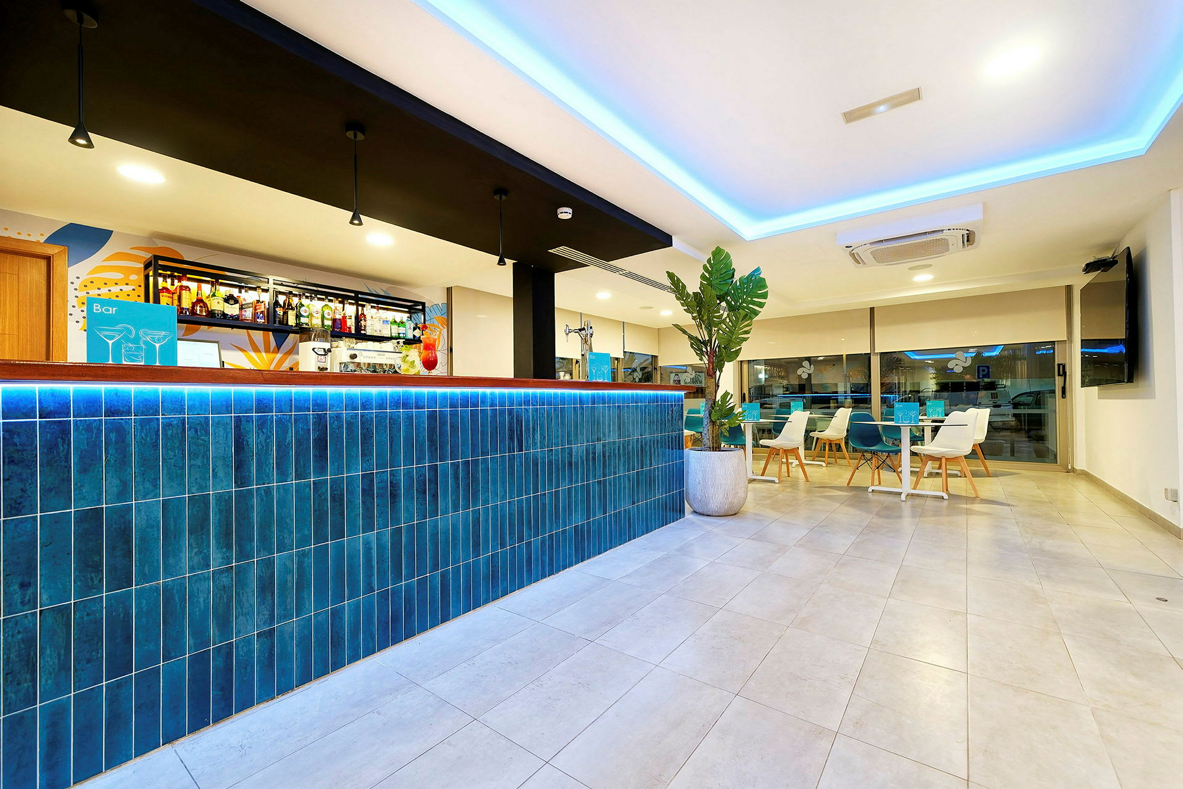 En moderne hotelbar med en blå flisebeklædt bardisk og stilrent interiør. I baggrunden ses siddeområder og en stor vinduesvæg. (AI-genereret tekst)