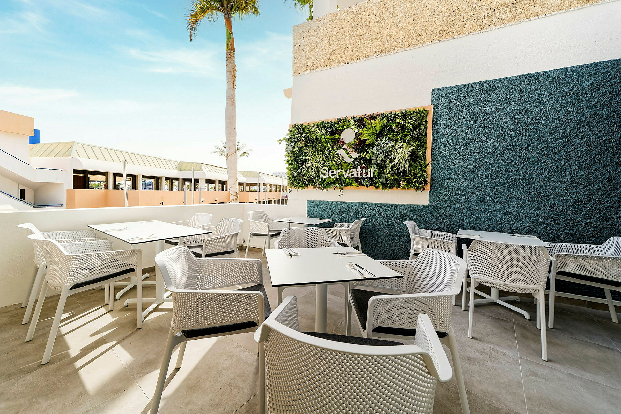 En udendørs terrasse med hvide borde og stole og dekorative planter på en væg. (AI-genereret tekst)