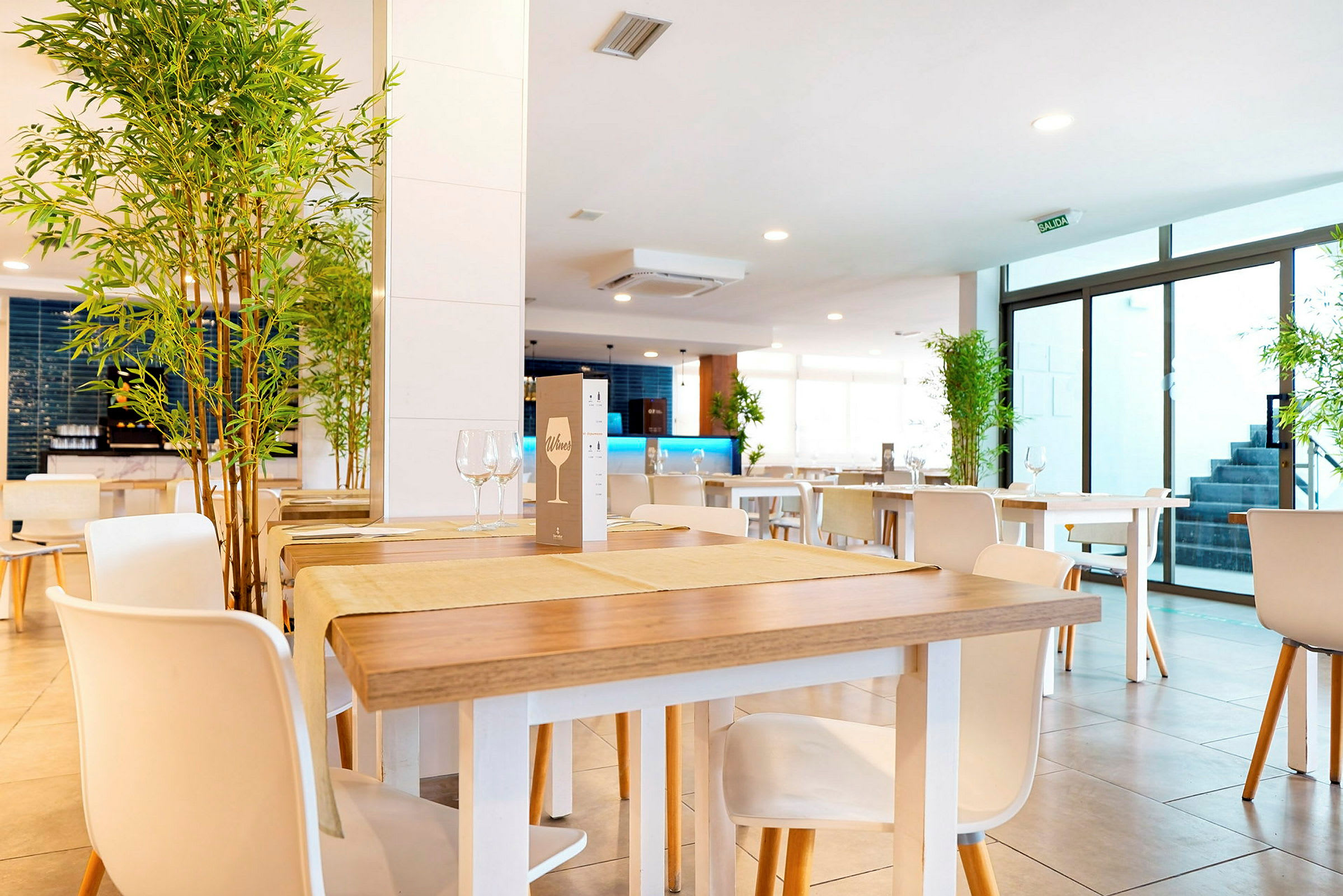 Et moderne og lyst indrettet restaurantområde med træborde og hvide stole samt dekorative planter placeret i hjørnerne. (AI-genereret tekst)