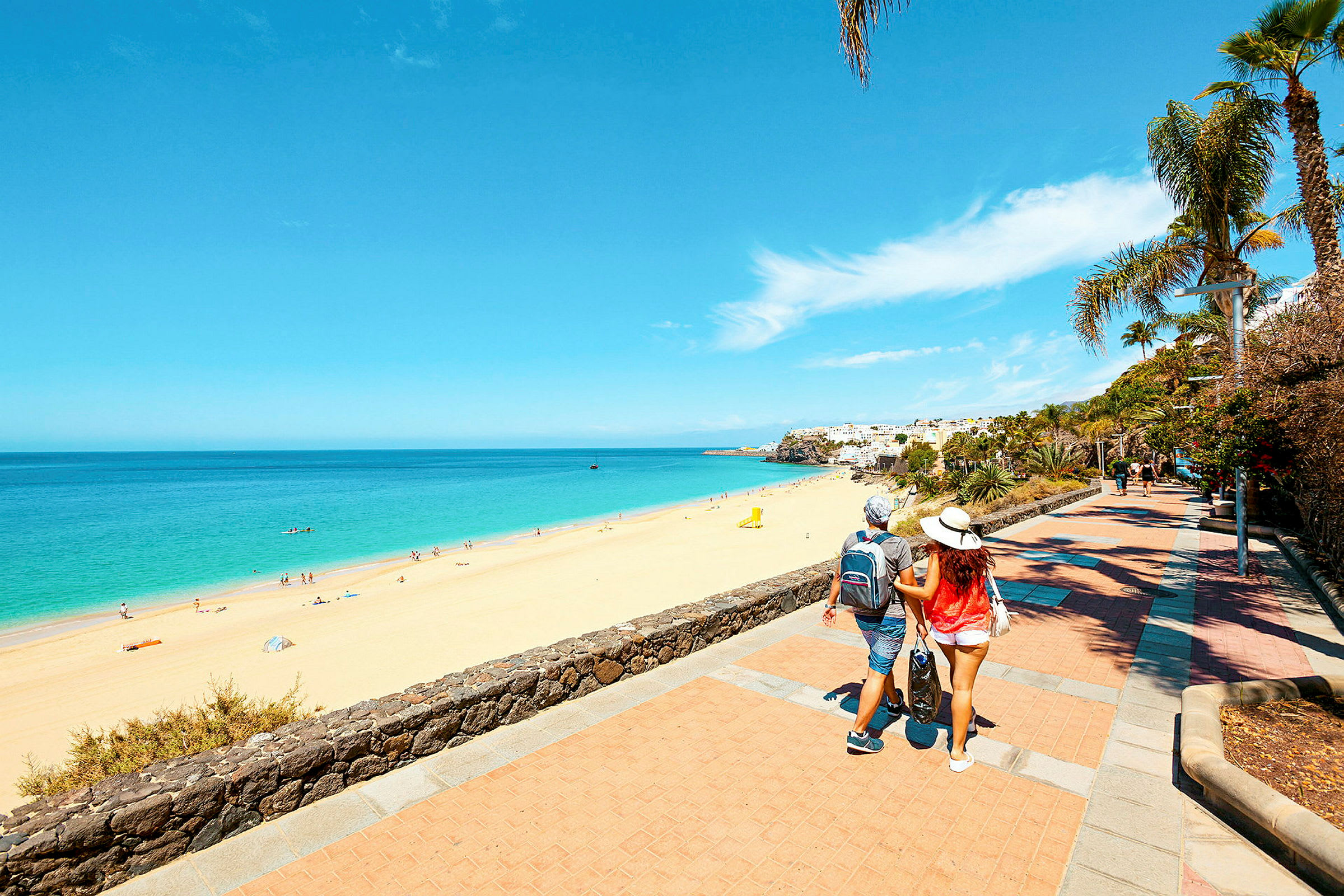 En strand med gyldent sand og klart blåt hav, omgivet af en promenade med palmer og anden vegetation. En person går langs promenaden. (AI-genereret tekst)