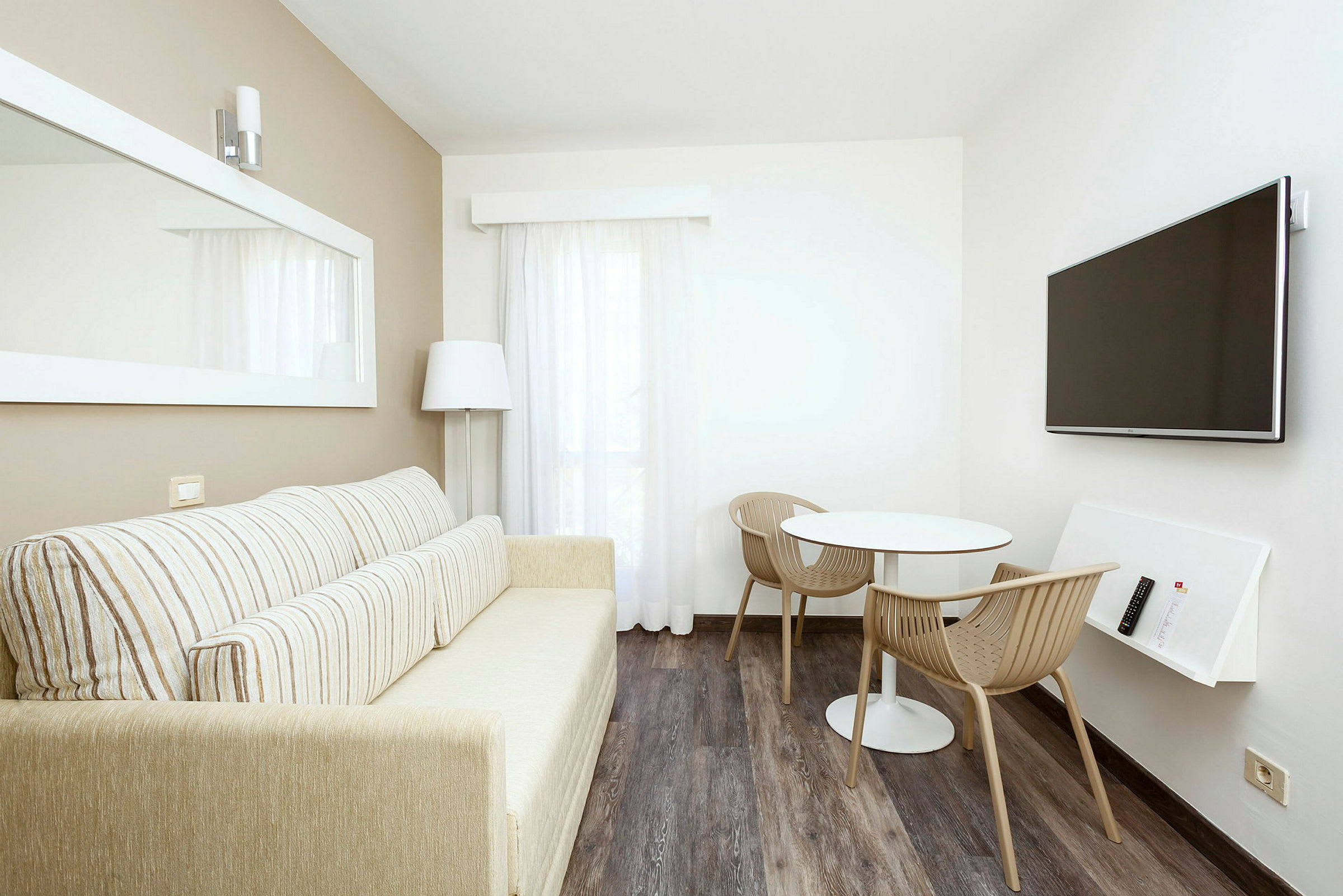 En lys stue med en sofa, et lille bord med to stole og et væghængt TV. (AI-genereret tekst)