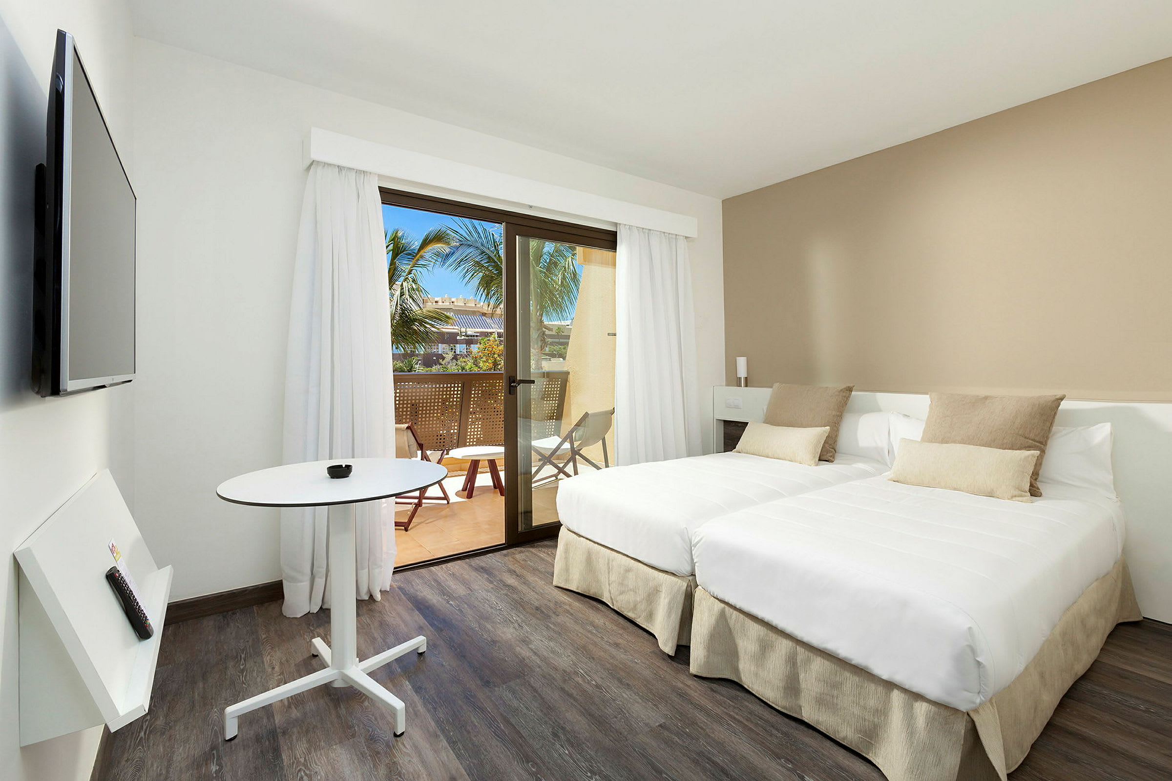 Et moderne hotelværelse med to enkeltsenge, et lille bord og et vægmonteret tv. Værelset har lyse farver og et vindue, der fører ud til en balkon med udsigt til palmer. (AI-genereret tekst)