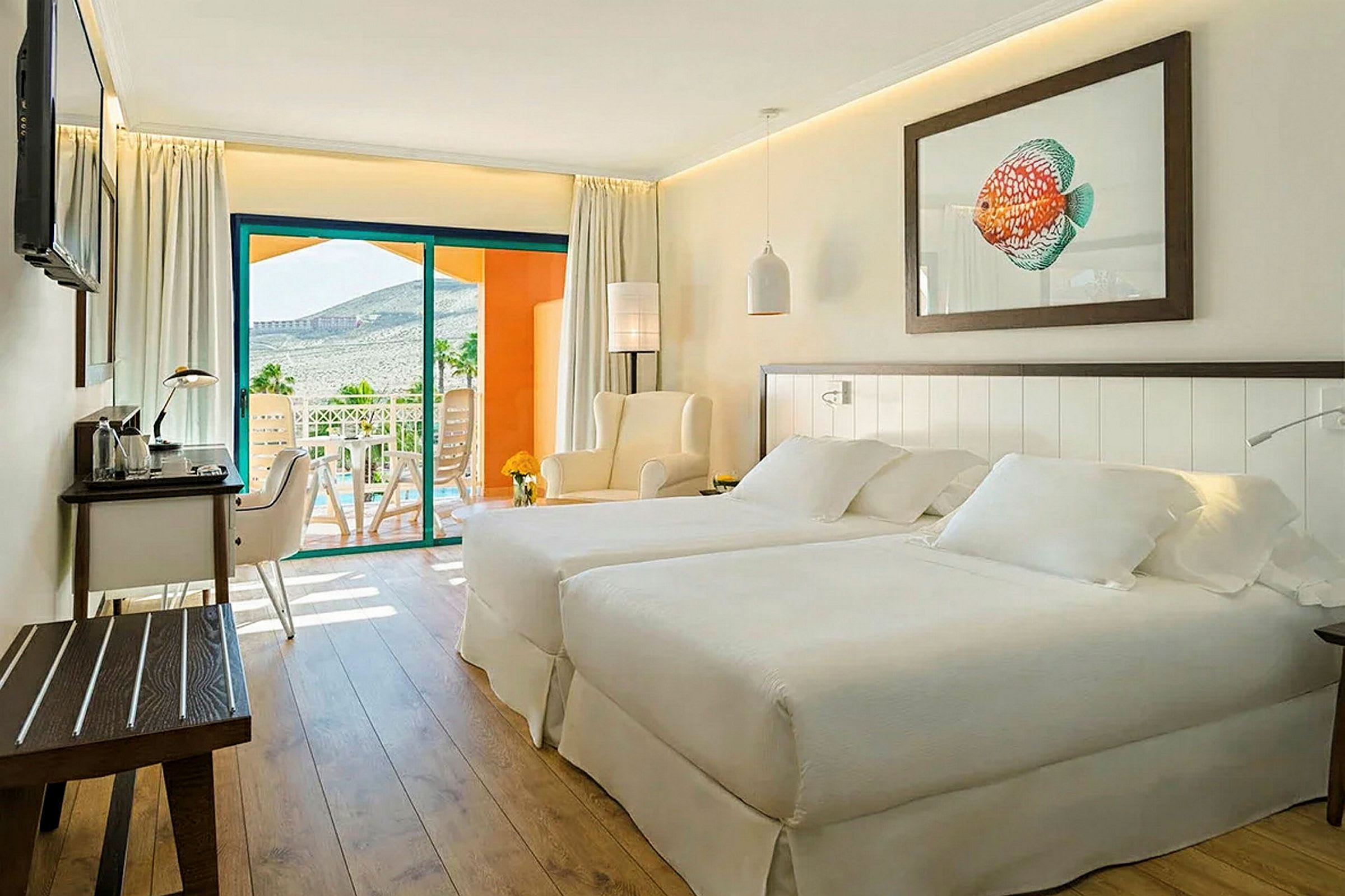 Et lyst og moderne hotelværelse med to senge, et siddeområde og et skrivebord. En balkon med udsigt over landskabet ses i baggrunden. (AI-genereret tekst)