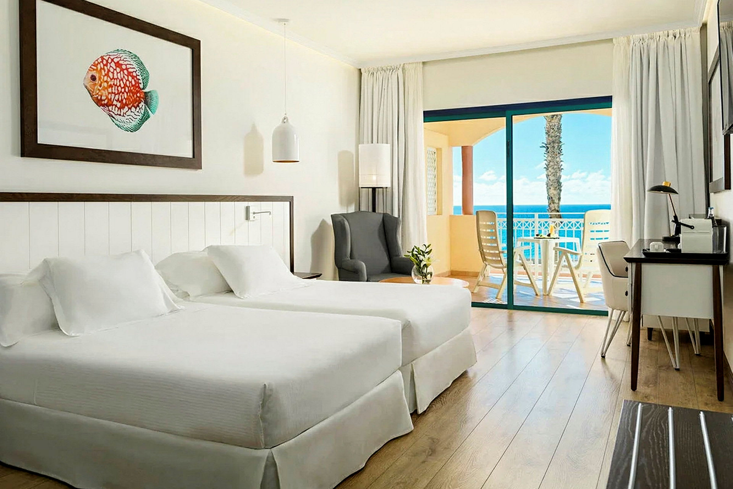 Et hotelværelse med to senge, en lænestol, et skrivebord og en balkon med udsigt til havet. (AI-genereret tekst)