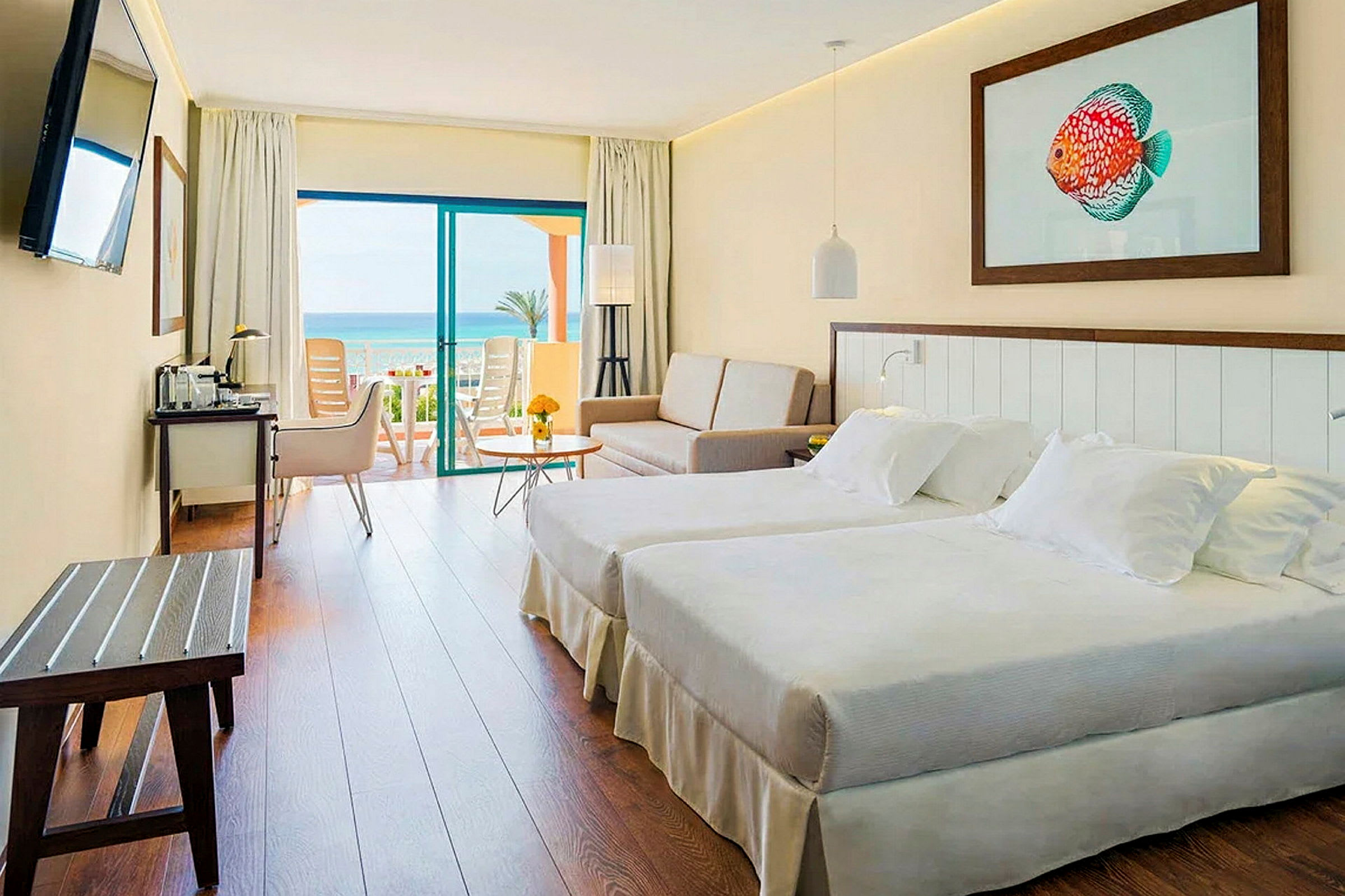 Et hotelværelse med to senge, en sofa og et bord. Værelset har en altandør, der fører ud til en altan med havudsigt. (AI-genereret tekst)