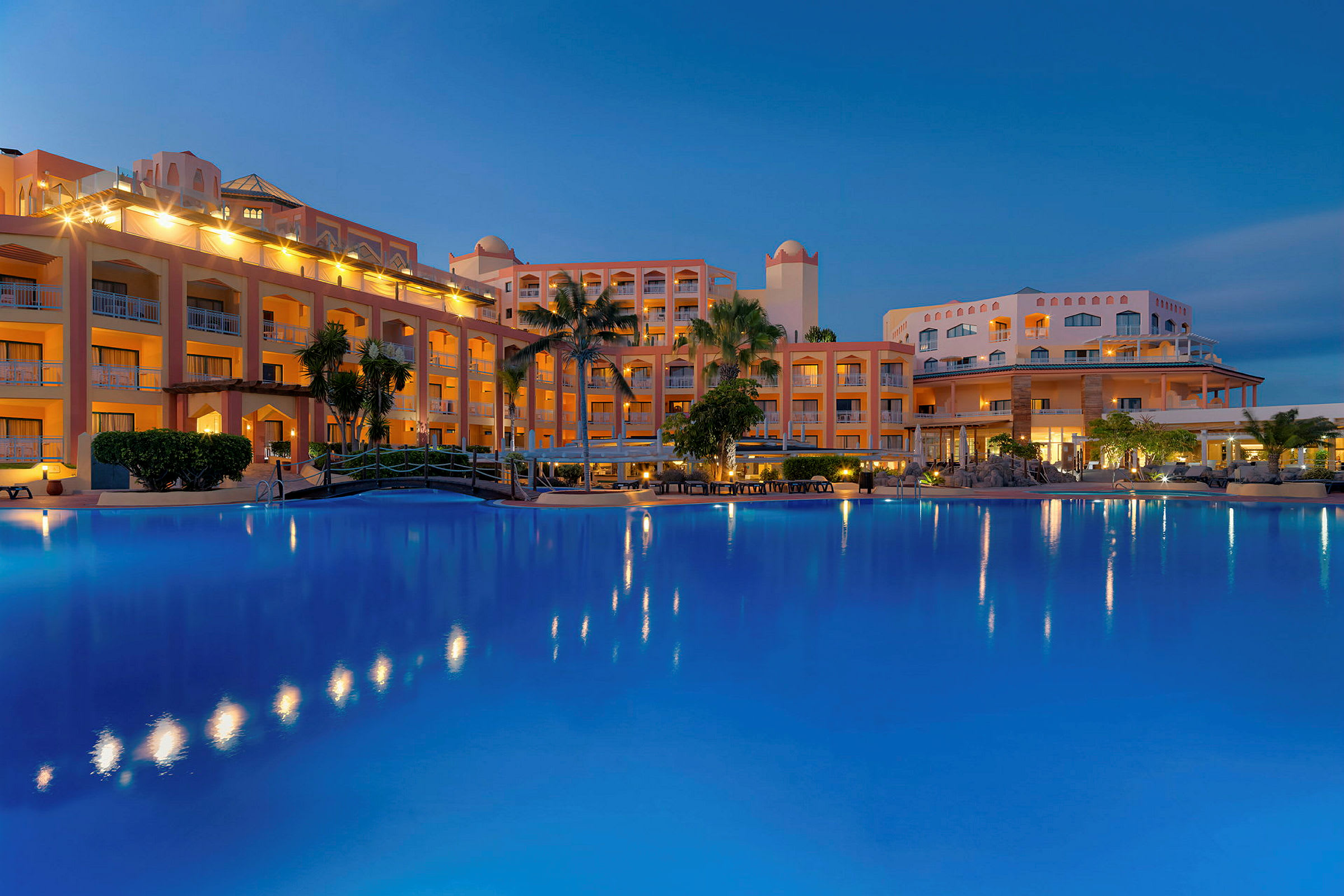 Et hotel ved tusmørke med en stor oplyst pool i forgrunden. (AI-genereret tekst)