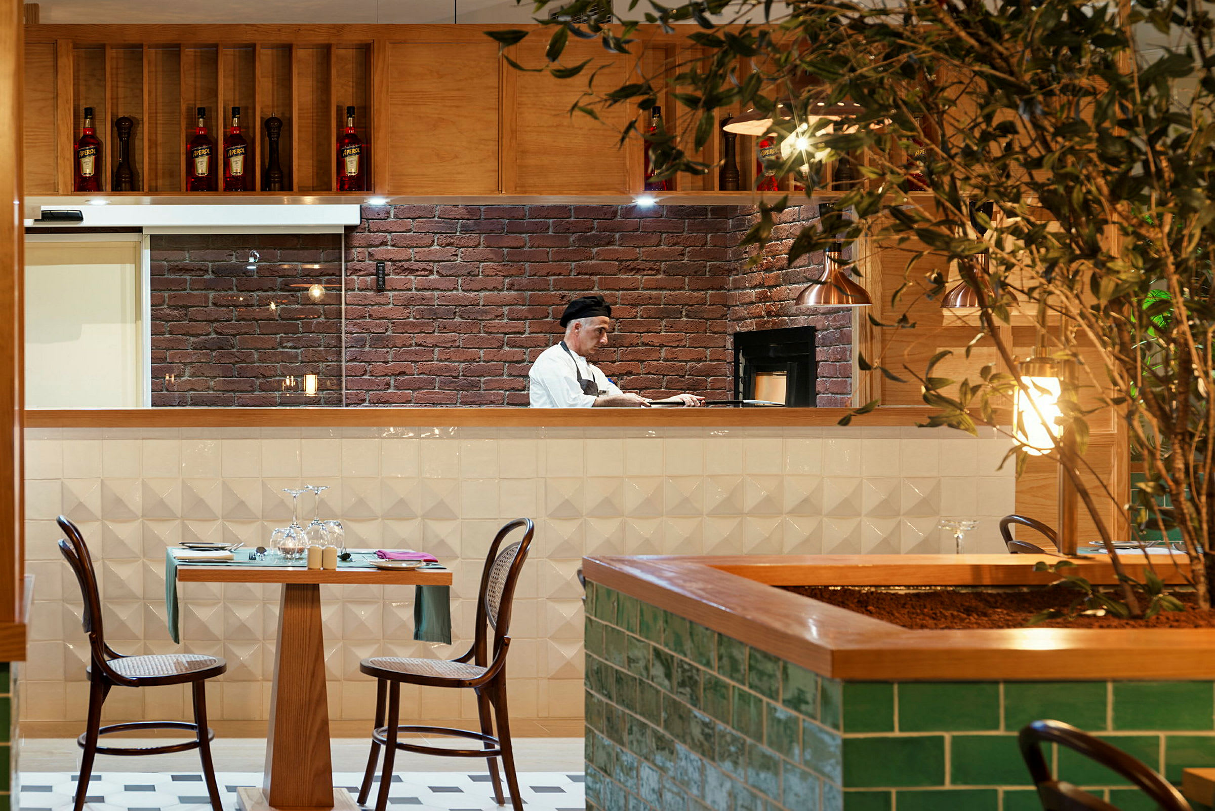 En restaurant med et åbent køkken, hvor en person arbejder. Et bord med to stole står foran køkkenet, og en plante er synlig i forgrunden. (AI-genereret tekst)