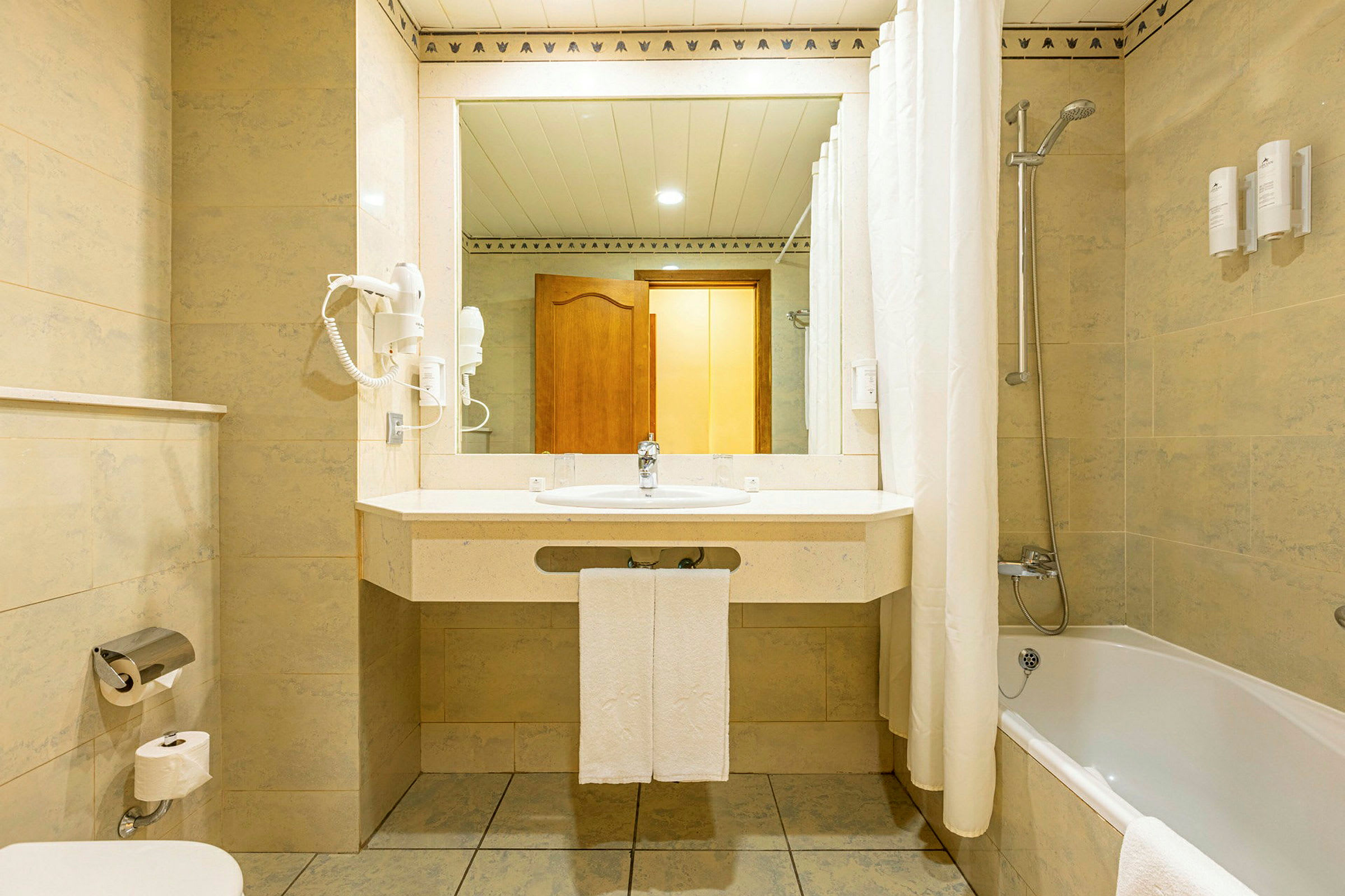 Et badeværelse med et spejl over en håndvask, toilet, badekar med bruser og lyse vægge. (AI-genereret tekst)