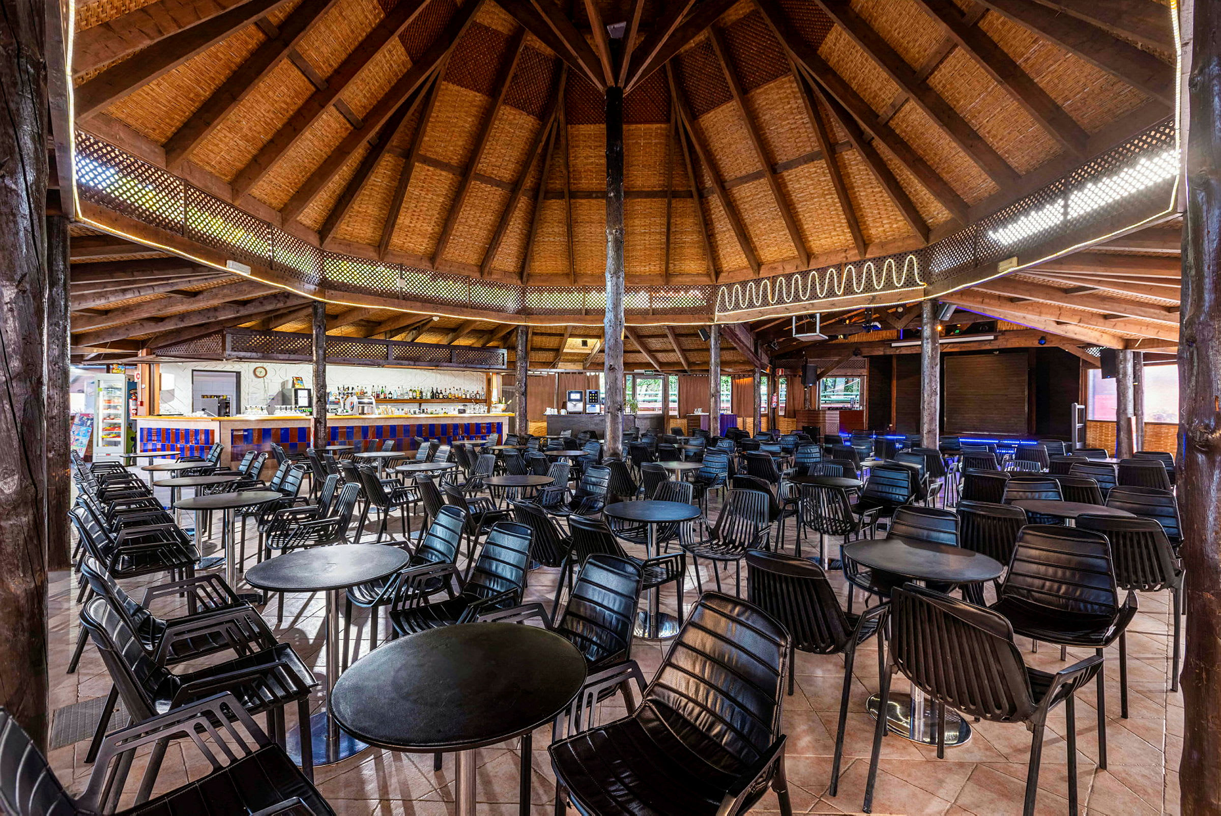 Et rundt hus af træ med stråtag indeholder en indrettet hal med borde og stole samt en bar i baggrunden. (AI-genereret tekst)