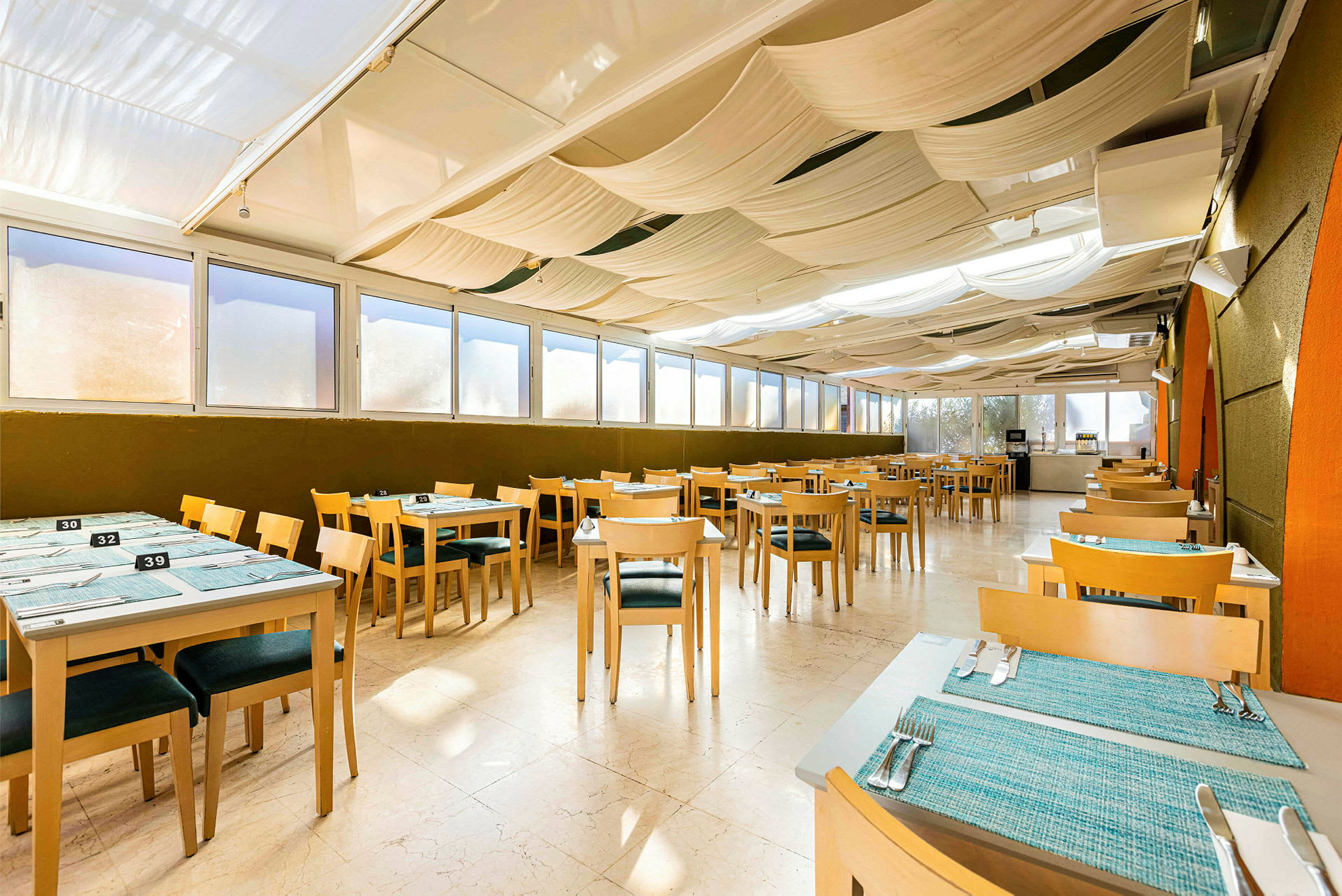 Billede af en indendørs restaurant med træborde og stole, blå bordmåtter og dagslys fra store vinduer. (AI-genereret tekst)
