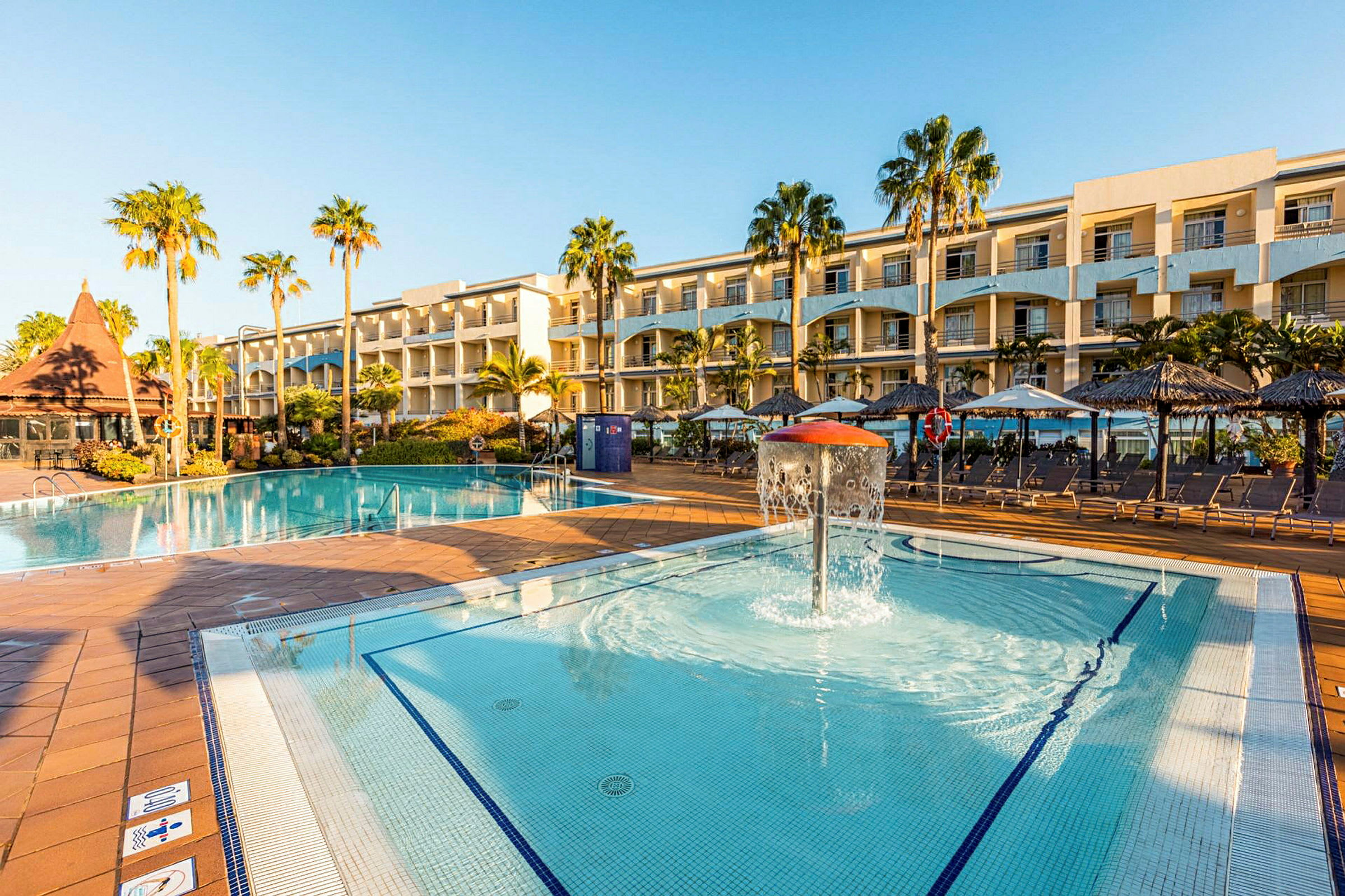 En stor hotelpool med omkringliggende liggestole og palmer, samt en mindre børnepool med springvand foran hotellets facade. (AI-genereret tekst)