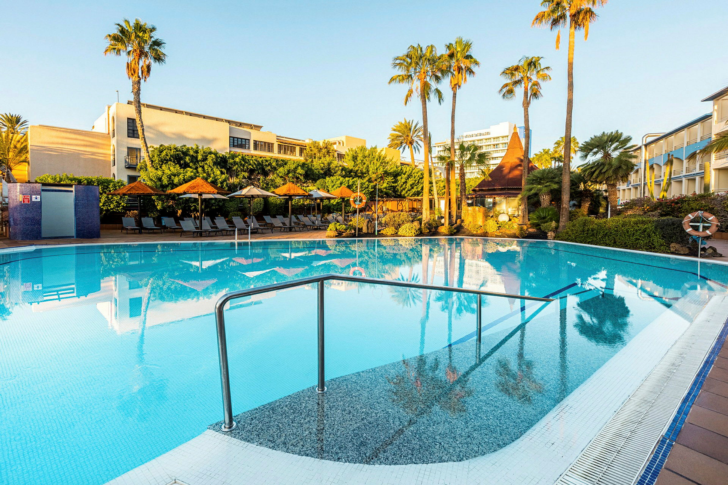 En udendørs swimmingpool omgivet af liggestole, parasoller og grønne palmer under en klar blå himmel. (AI-genereret tekst)