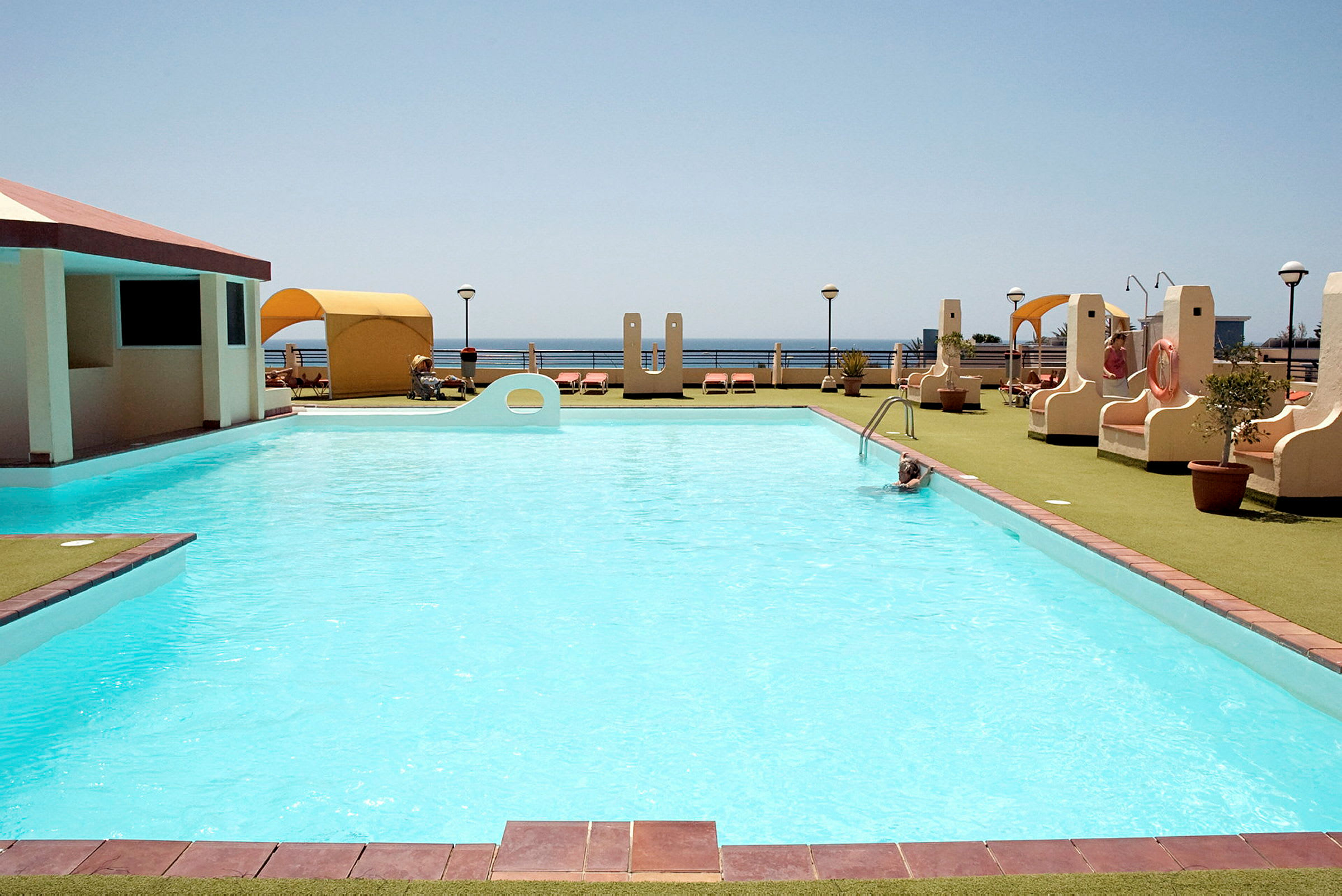 Billede af en udendørs pool omgivet af liggestole og bungalower, med havet i baggrunden. (AI-genereret tekst)