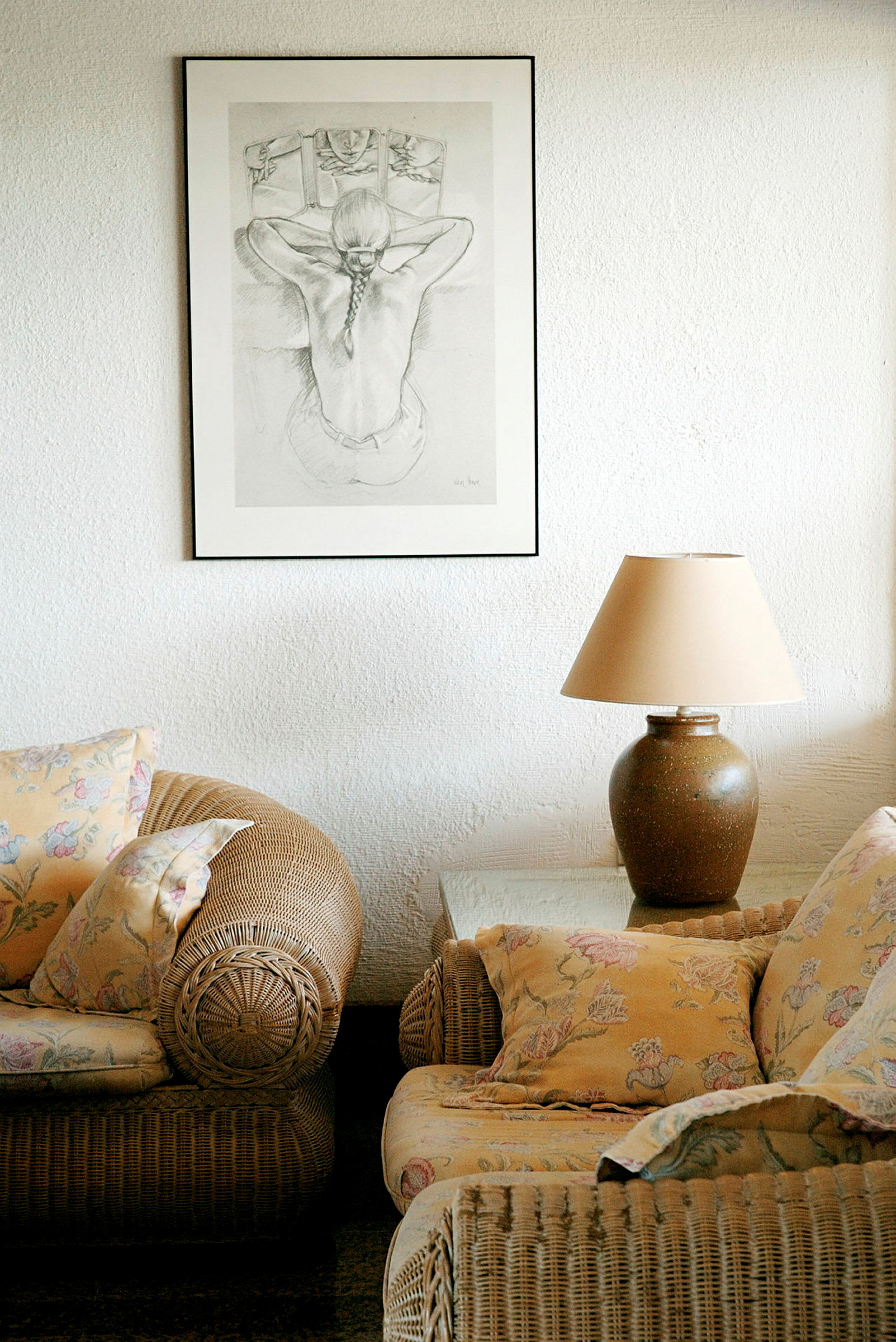 Billede af en stueinteriør med et billede på væggen, en lampe på et bord og rottingmøbler med blomstrede puder. (AI-genereret tekst)